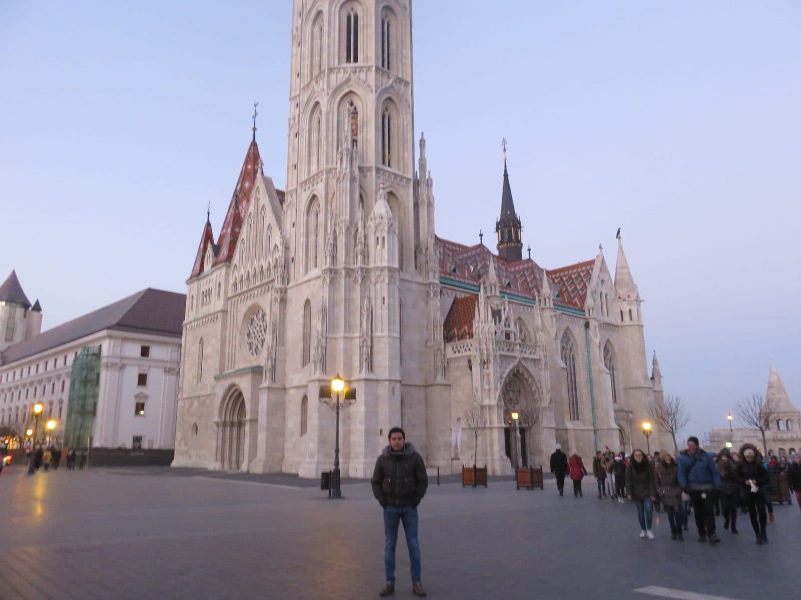 Matthias Church in Budapest at dusk on a winter afternoon, its diamond-patterned tiled roof in red, green and white below a tall pale gothic tower, street lamps already lit across the empty square, a man in a winter coat standing in the foreground, and a long file of people walking past on the right with a white turret of Fisherman's Bastion beyond them
