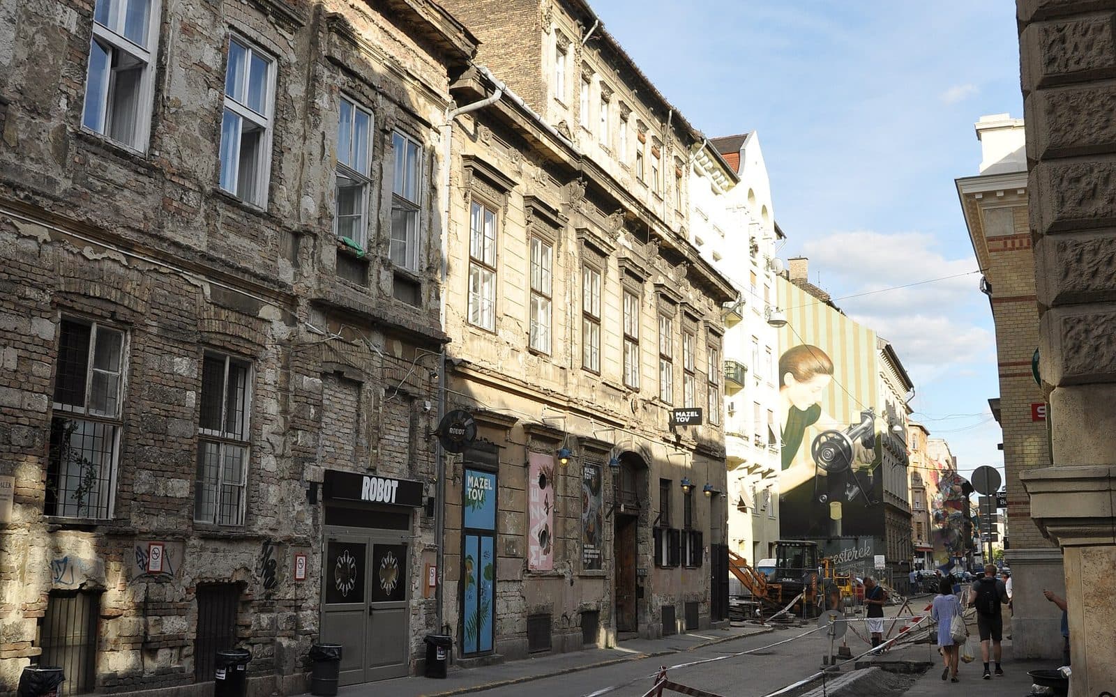 Akacfa utca in the Budapest Jewish Quarter, with a black hanging MAZEL TOV sign and painted Mazel Tov panels on a building whose stucco has fallen away to bare brick, the ROBOT bar next door, and a large mural of a woman at a sewing machine further down the street