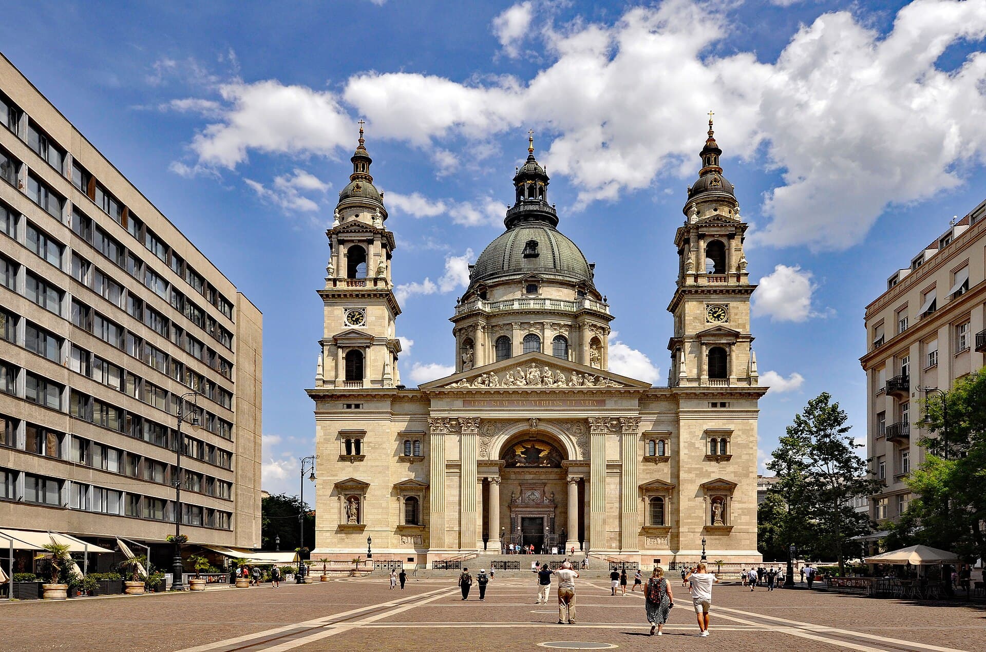 The west front of St Stephen's Basilica in Budapest on a sunny day: a pale stone neoclassical facade with a columned portico and sculpted pediment, flanked by two bell towers with clock faces, and the green copper dome rising behind, with people crossing the paved square in front