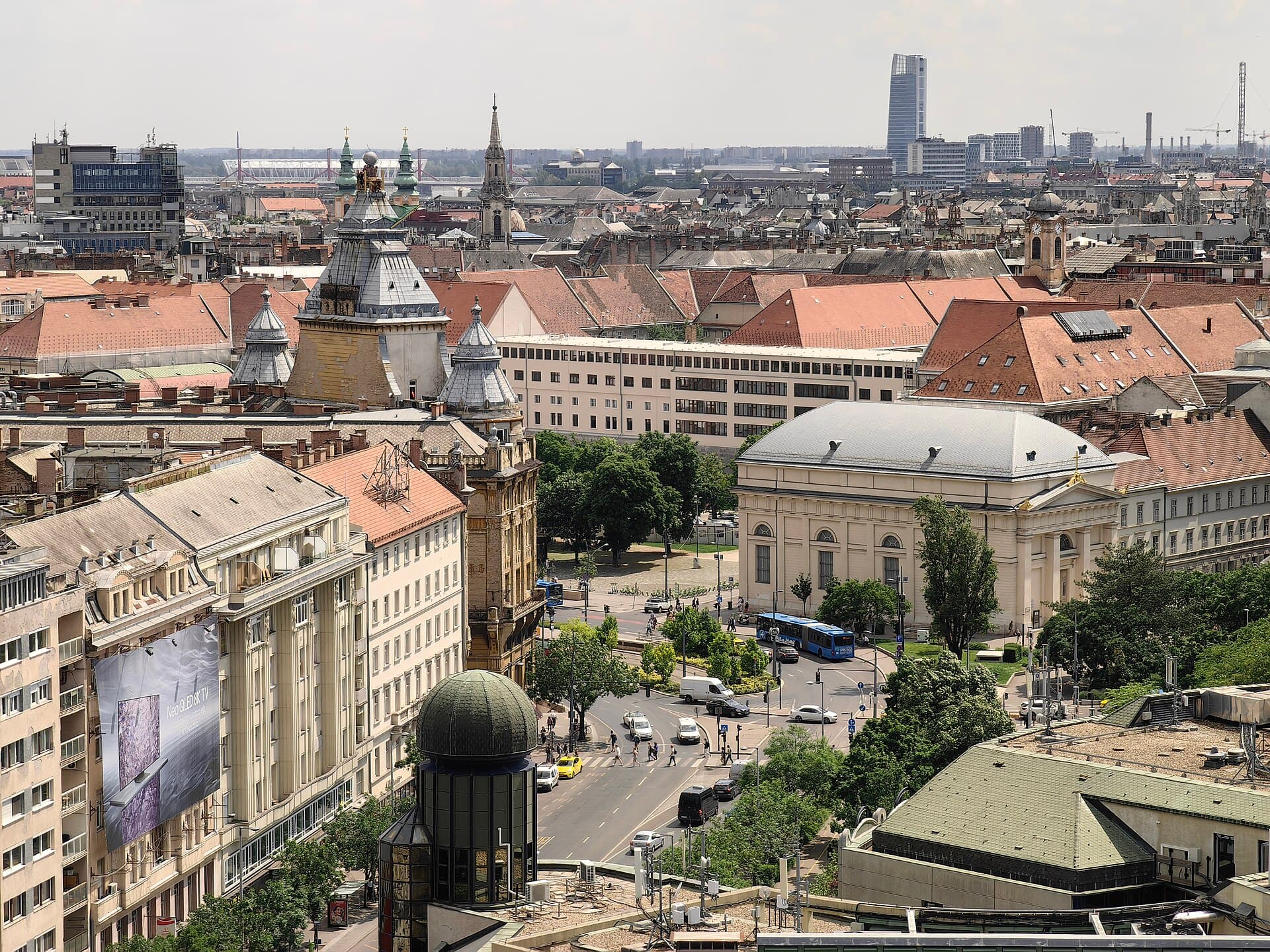 The view from the panoramic terrace of St Stephen's Basilica looking out over Pest: a dense field of red-tiled and grey rooftops, a leafy square with a neoclassical building and a blue city bus, a large advertising hoarding on a building wall in the foreground, and a modern glass tower on the hazy horizon