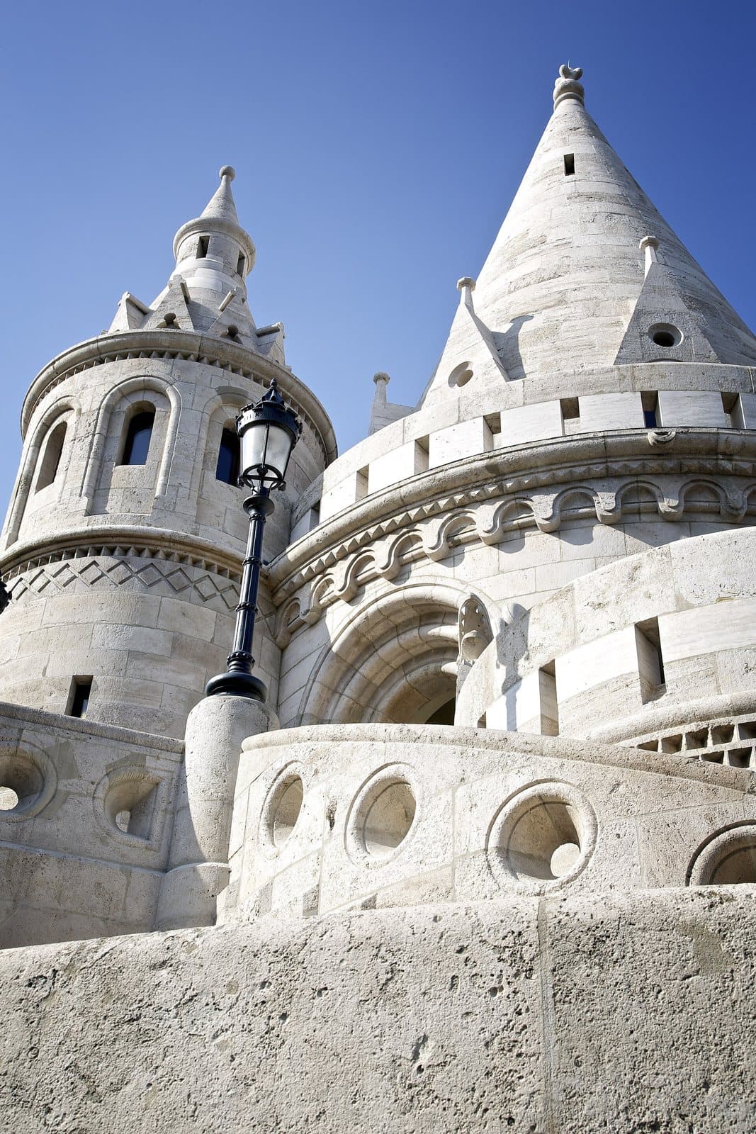 Two of the white stone turrets of Fisherman's Bastion photographed from below against a deep blue sky, showing their conical roofs, arched openings and the carved zigzag and circular decoration of the parapet, with an ornate dark lamp post between them