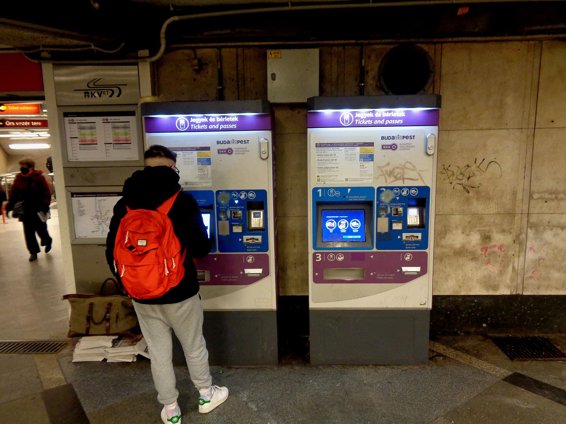 Two purple and white BKK ticket machines labelled Jegyek és bérletek, Tickets and passes, in a Budapest metro underpass, with a traveller in a red backpack using the left-hand one