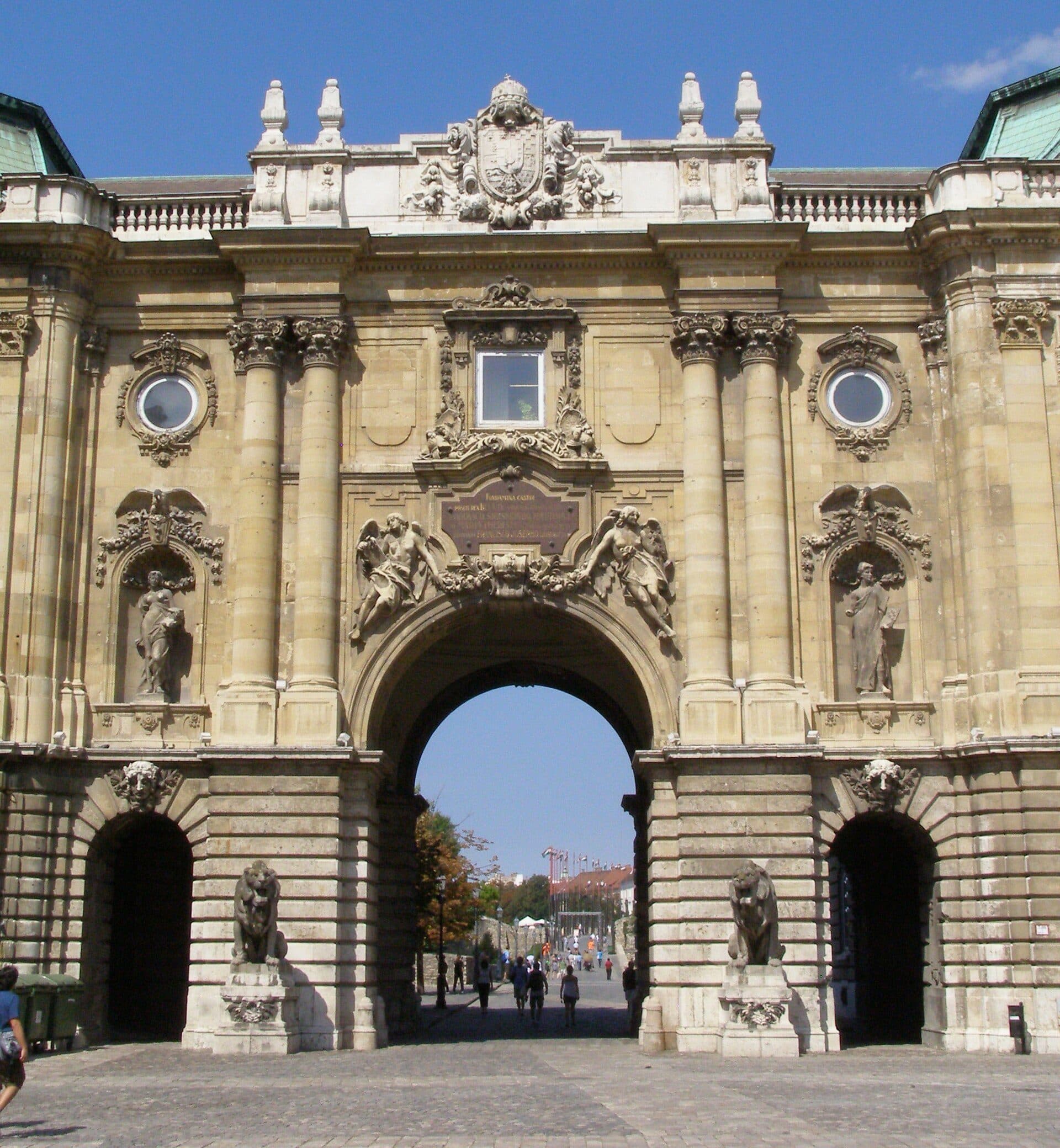 The baroque gateway to the Lion Courtyard at Buda Castle, with a stone lion on each side of the central arch, the Hungarian coat of arms above it, and people walking through the archway