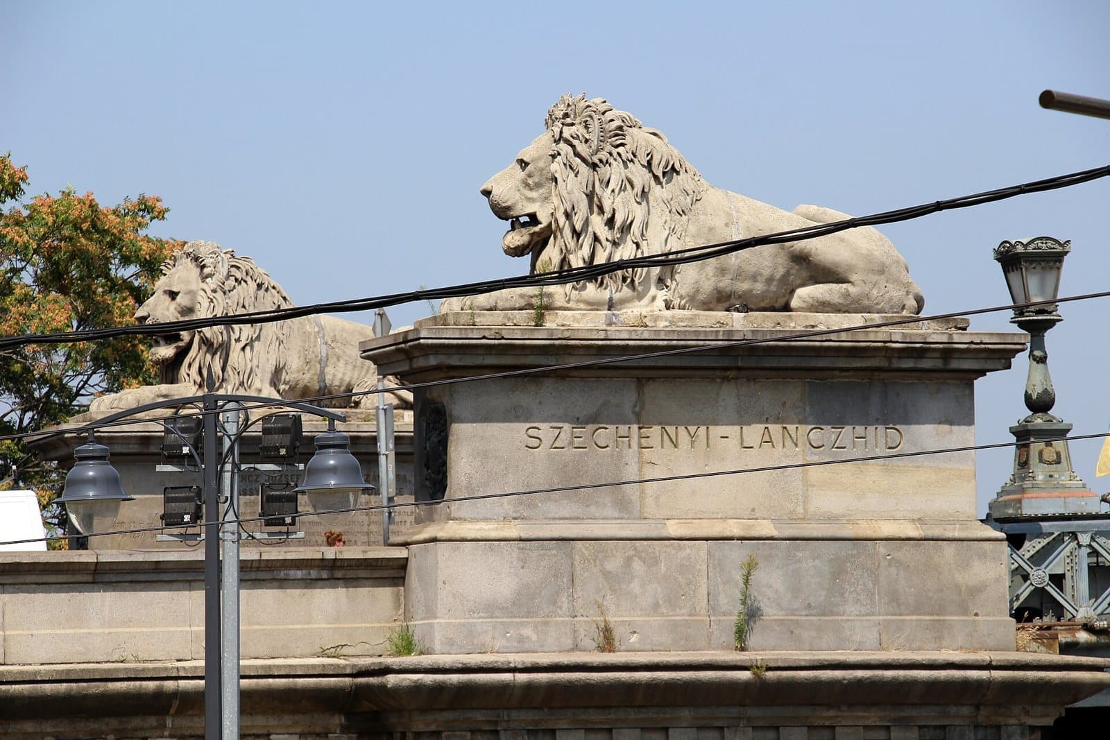Two carved stone lions lying on their plinths at the end of the Chain Bridge in Budapest, seen from below against a pale sky, with SZECHENYI-LANCZHID carved into the stone of the nearer plinth and tram wires crossing the view
