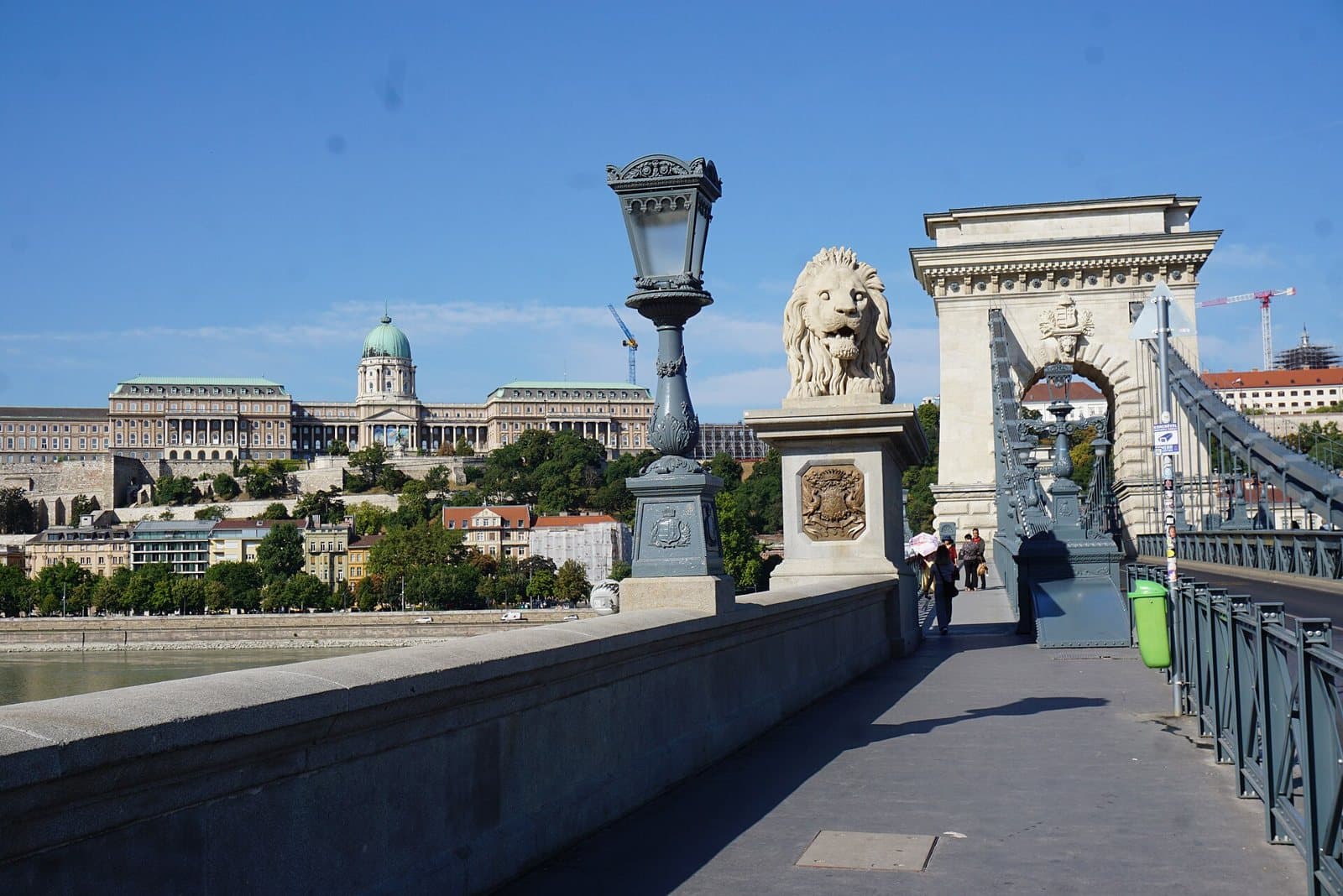 Walking across the Chain Bridge in Budapest: a wide stone pavement leading towards the bridge's pale stone tower arch, with a carved lion on a plinth and an ornate lamp post beside it, a few pedestrians further along, an empty carriageway to the right, and Buda Castle on the hill across the Danube