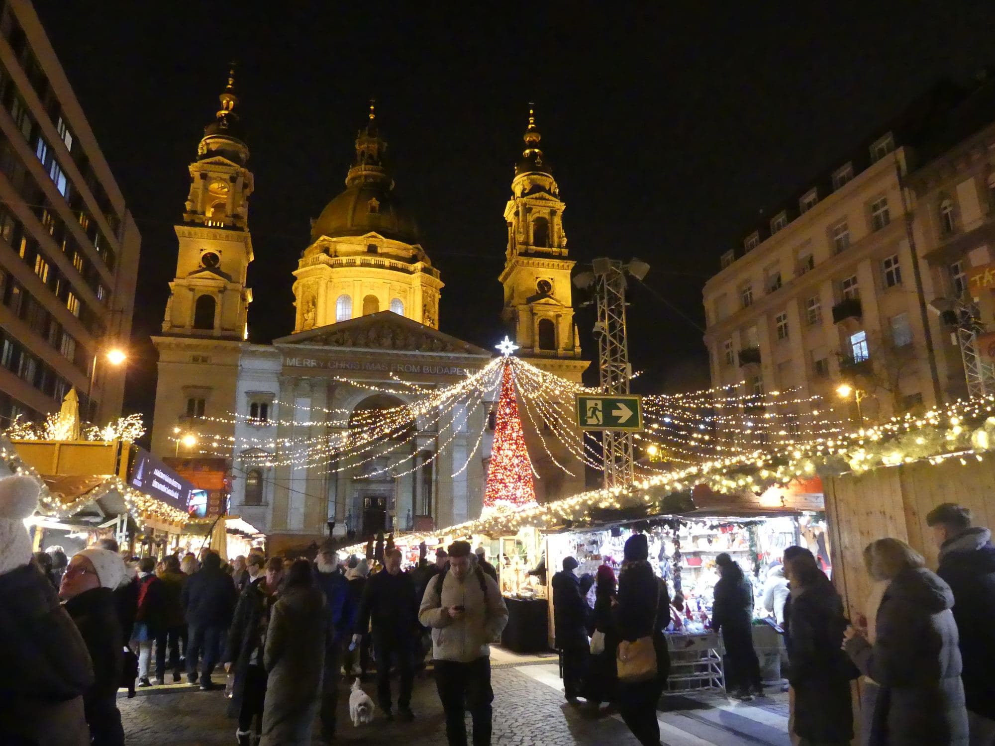 El mercado de Navidad de la plaza de la Basílica de San Esteban de noche, con la fachada iluminada en dorado y el rótulo MERRY CHRISTMAS FROM BUDAPEST, un árbol cónico de luces rojas, toldos de guirnaldas cruzando la plaza, casetas de madera a los lados y una multitud abrigada