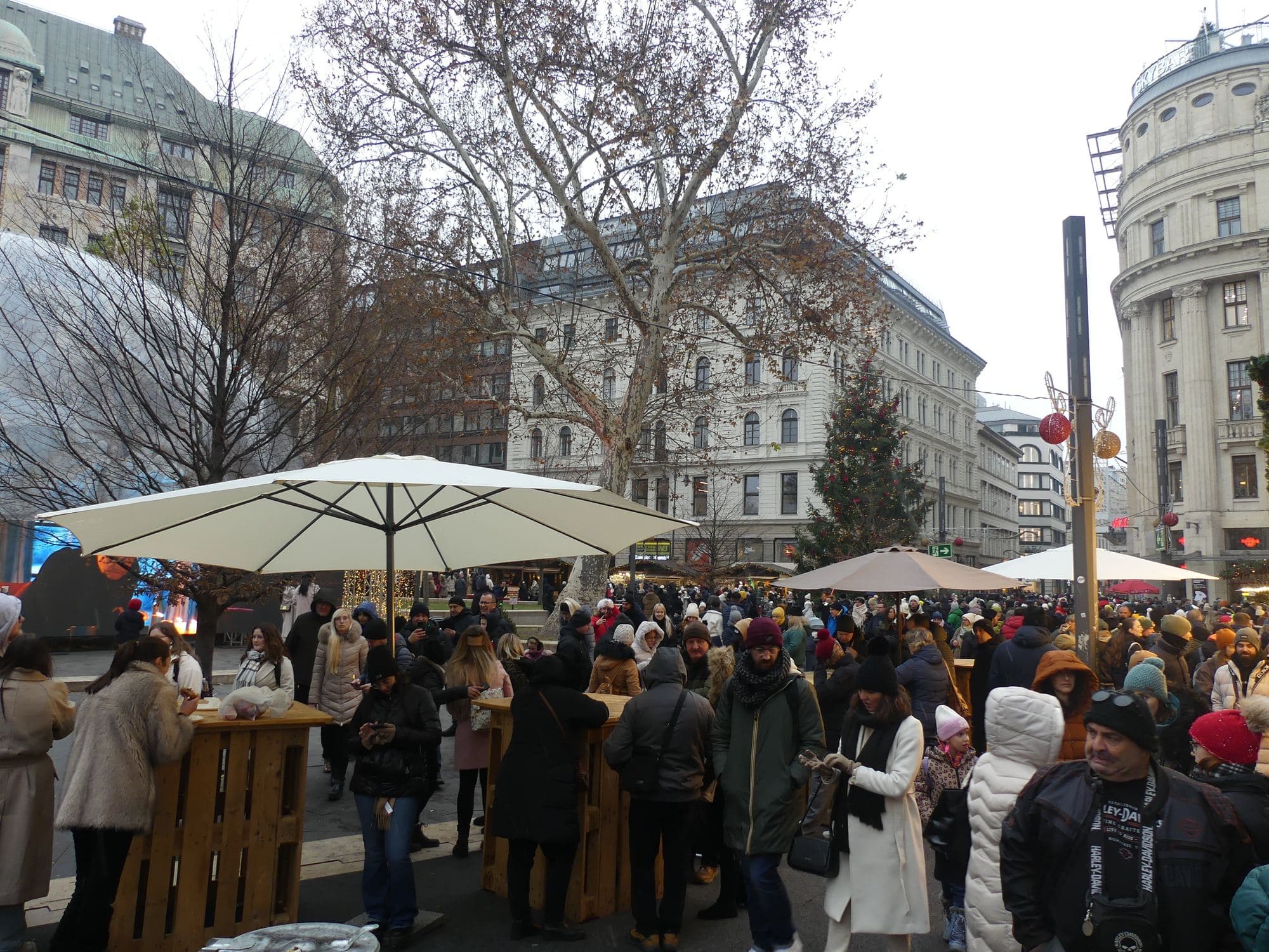 The Vorosmarty ter Christmas market in Budapest packed with people, with standing tables, parasols and a decorated Christmas tree on the square