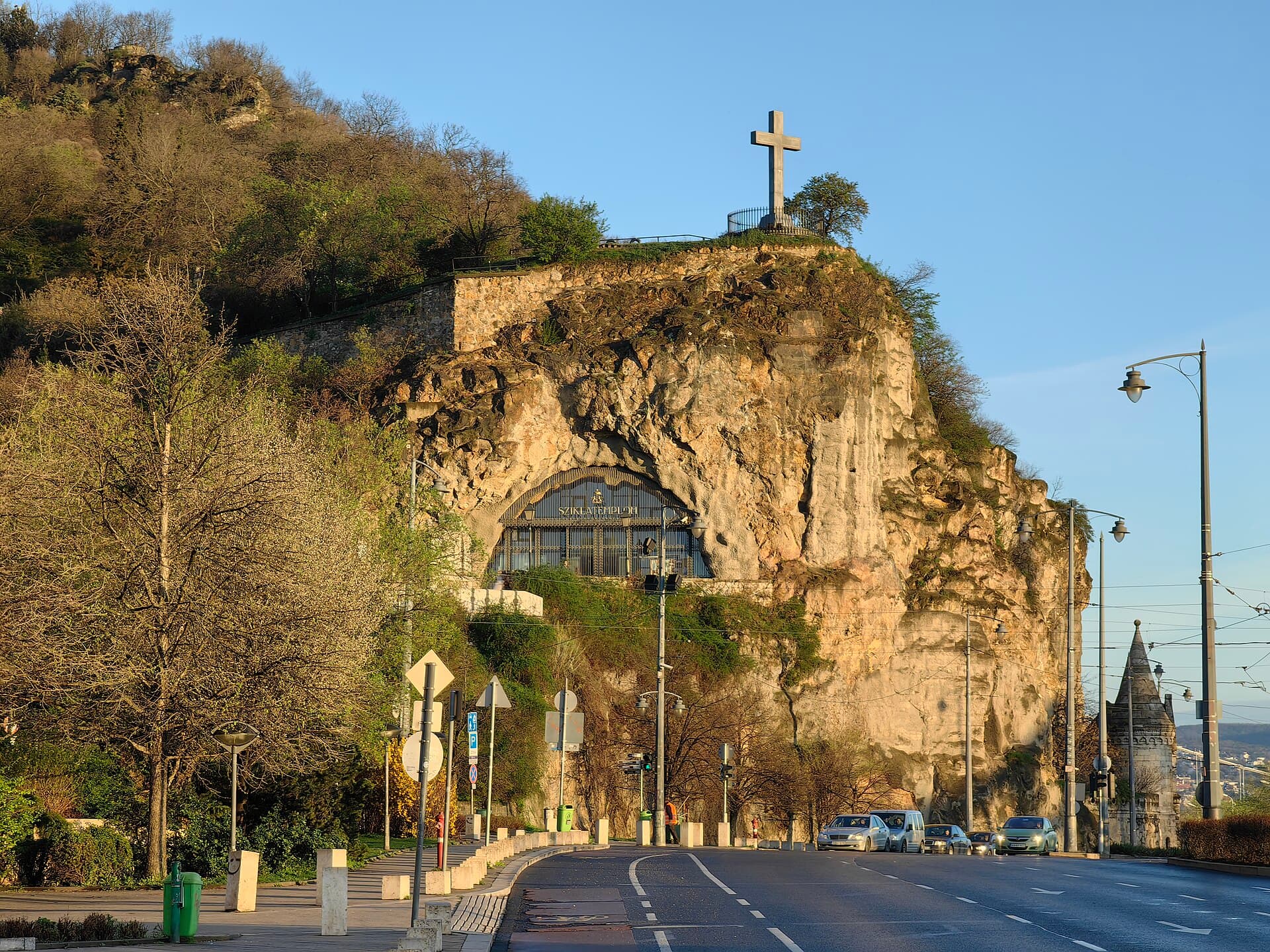 The entrance to the Cave Church in the cliff face at the foot of Gellert Hill, an arched iron grille set into the rock under a sign reading SZIKLATEMPLOM, with a large stone cross on the clifftop above and the embankment road in front