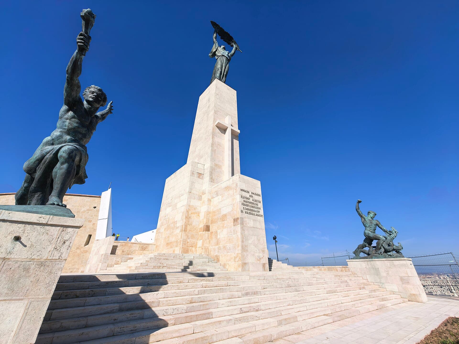 The Liberty Statue on Gellert Hill seen from below against a clear blue sky, with two bronze subsidiary figures either side, standing on new pale stonework and broad new steps, with construction hoarding to the left and site fencing to the right