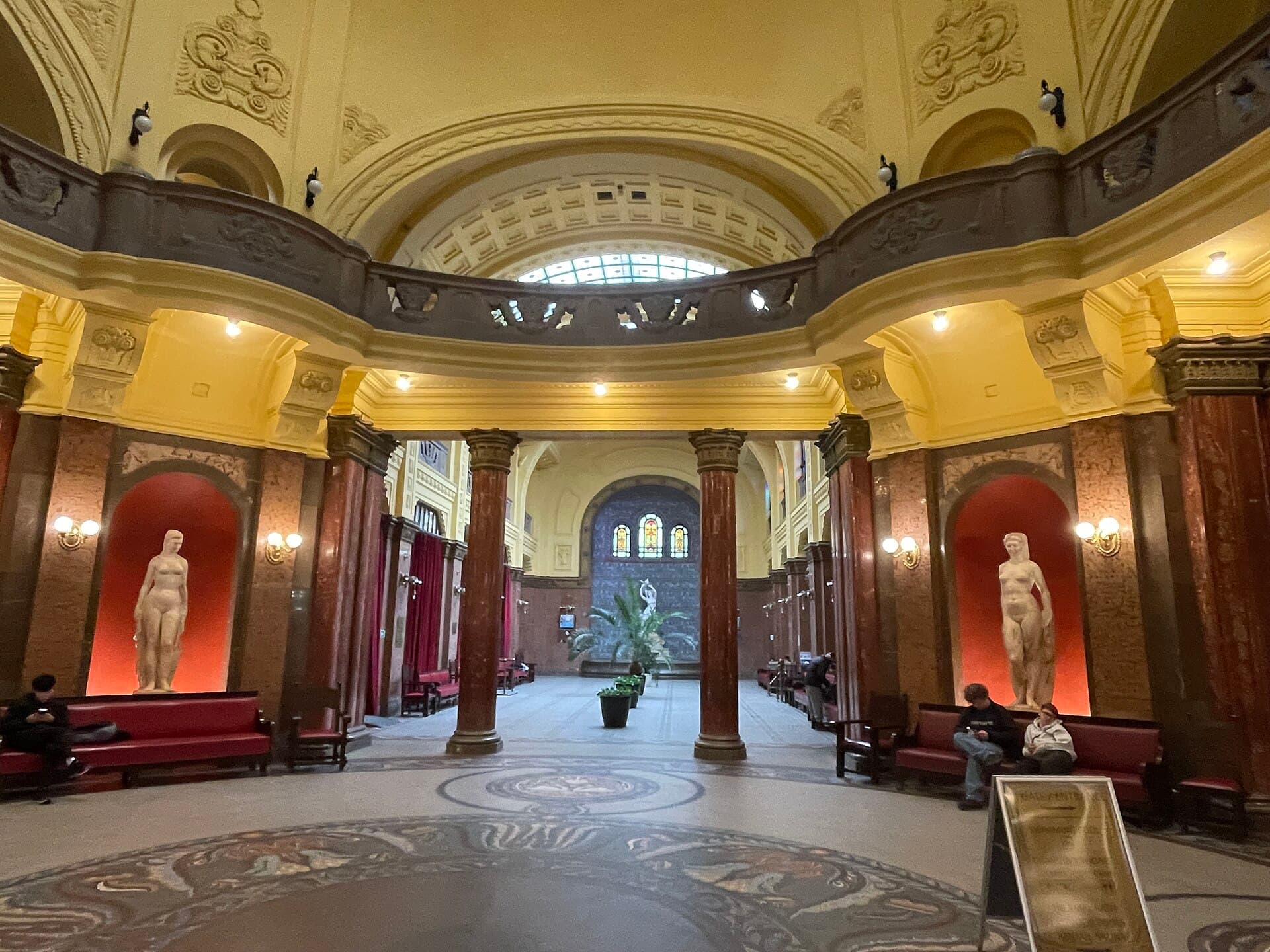 The domed entrance lobby of Gellert Baths in April 2024, with red marble columns, two pale stone nude statues lit in arched red niches, red benches where a handful of people sit waiting, a mosaic floor and a stained-glass window in the far hall