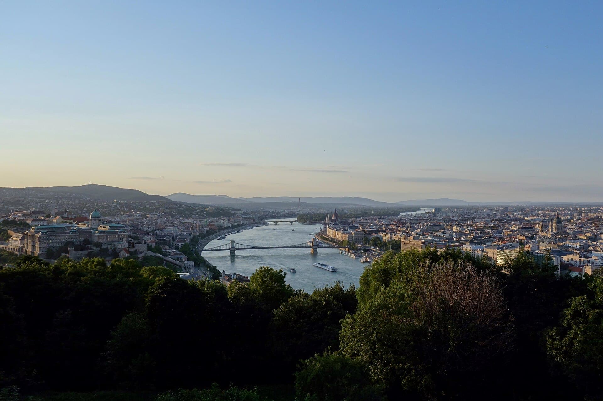 The view north from Gellert Hill at dusk: Buda Castle on the left bank, the Chain Bridge crossing the Danube, the Parliament dome further up the far bank and the Basilica dome on the right, with dark treetops filling the foreground