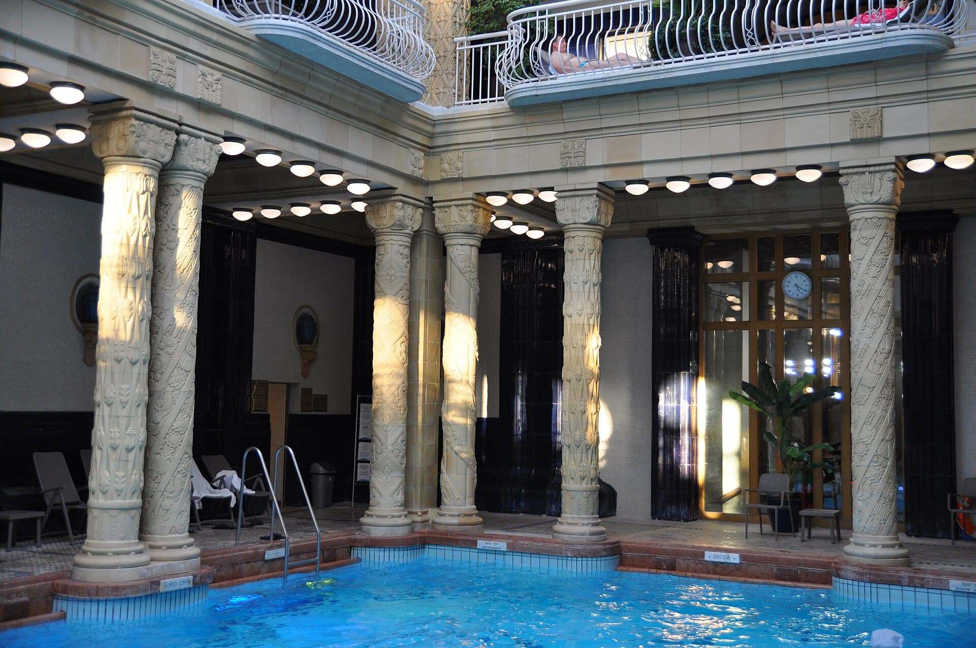 The Art Nouveau indoor swimming hall at Gellert Baths, with carved cream columns wound with sculpted ribbon, black glazed tiles behind them, a ring of round ceiling lamps, a first-floor gallery where two people are sunbathing, and depth markers reading 180 CM and 200 CM at the pool edge