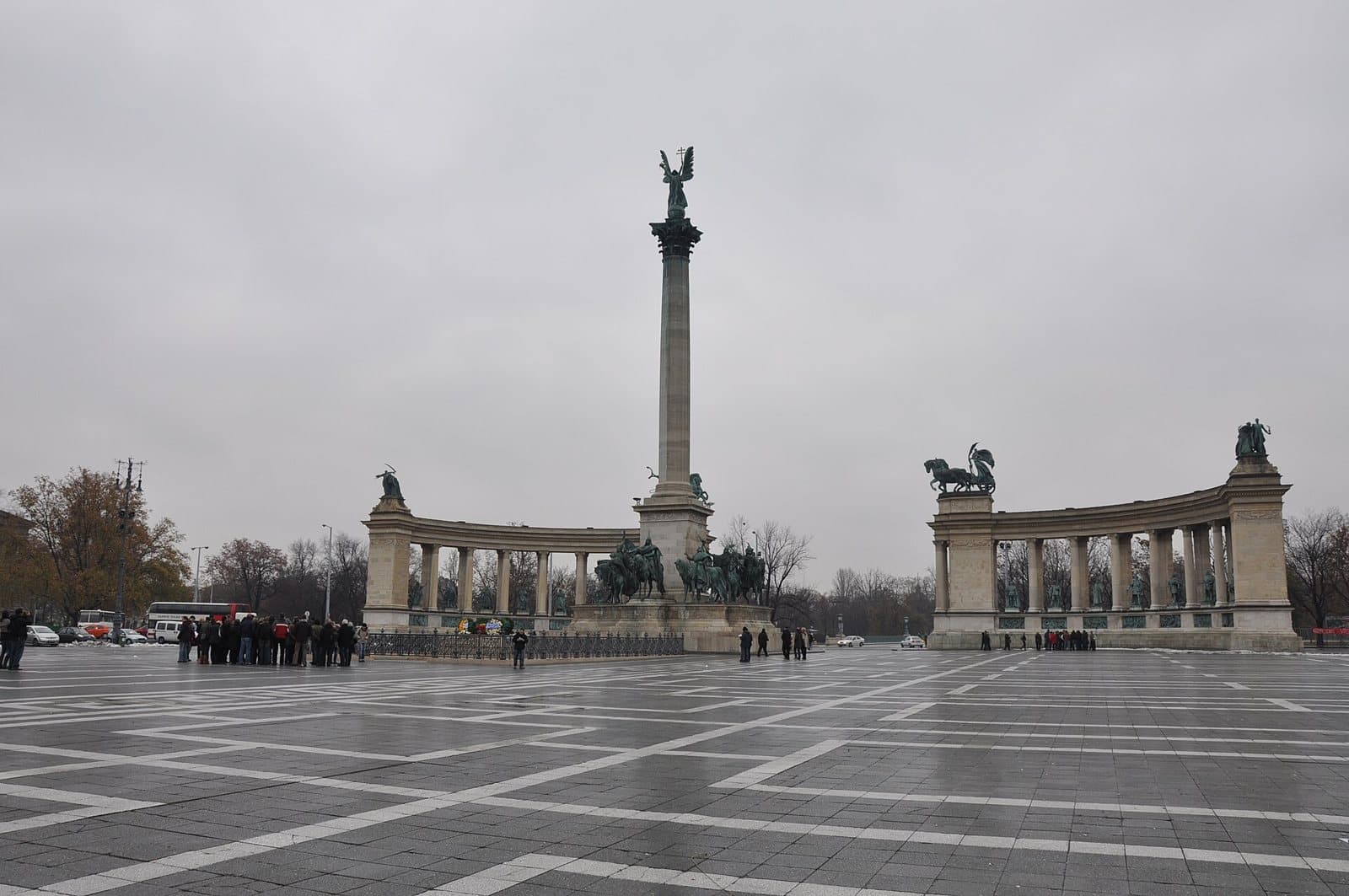 Heroes' Square in Budapest on an overcast day: a wide wet paved square with a tall stone column topped by a winged statue of the Archangel Gabriel, a bronze group of mounted chieftains at its base, and two curved colonnades of columns holding statues on either side, with a tour group standing at the monument