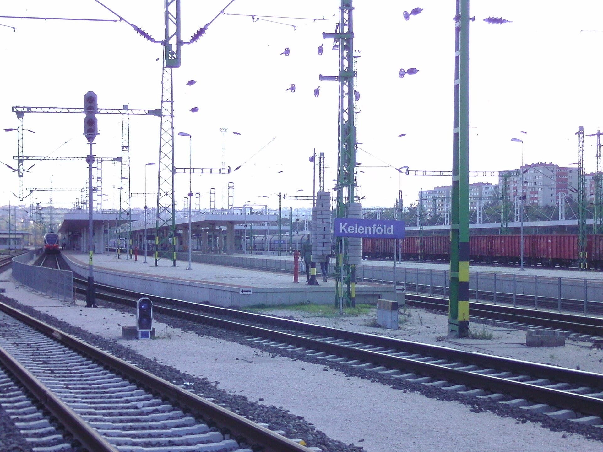 Andenes y catenarias en la estación de Budapest-Kelenföld, con un cartel azul con el nombre Kelenföld y vagones de mercancías en las vías del fondo