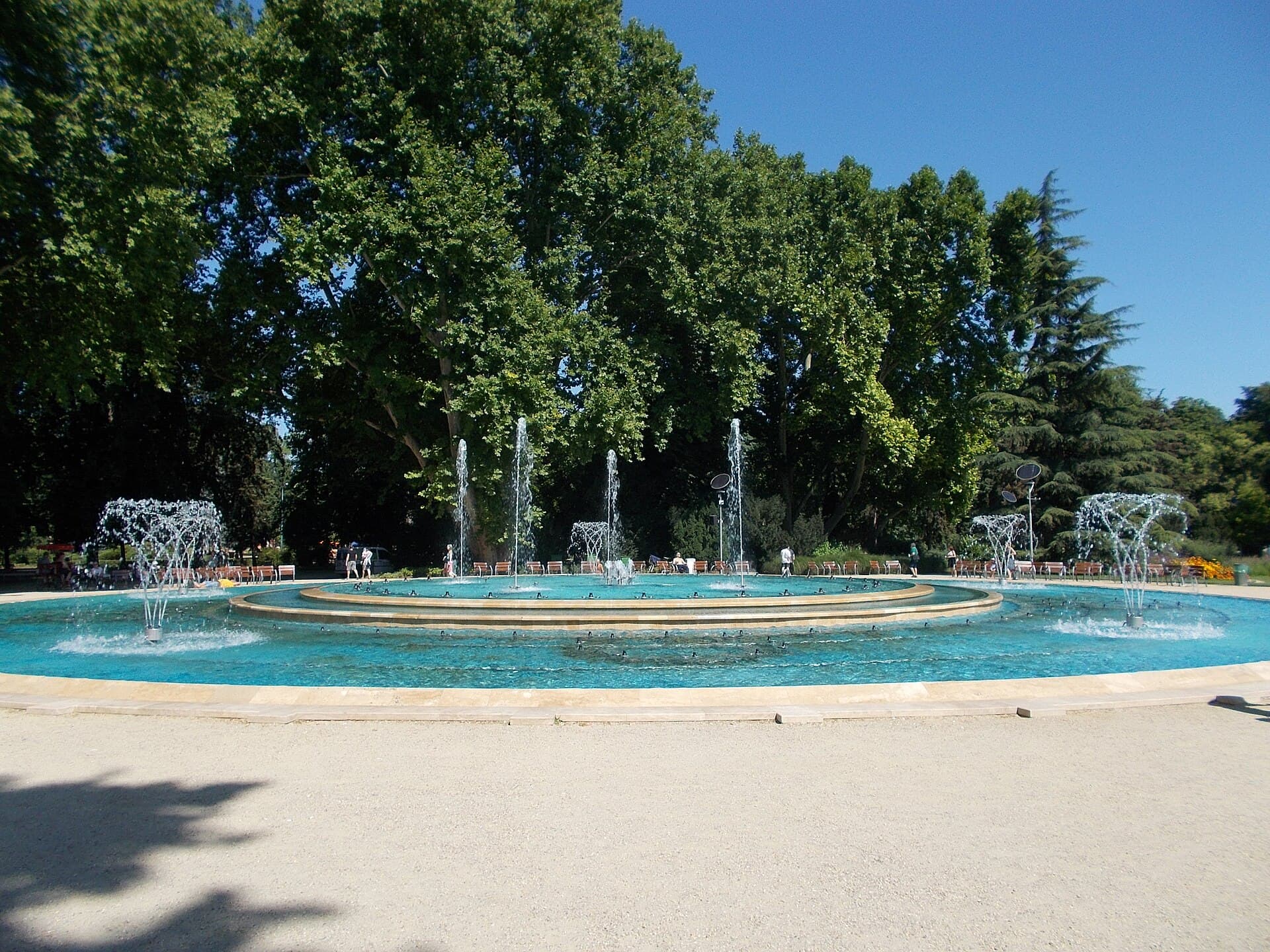 The musical fountain on Margaret Island seen from the gravel path: a wide circular basin with concentric stone steps, rows of water jets playing, park benches and tall plane trees behind