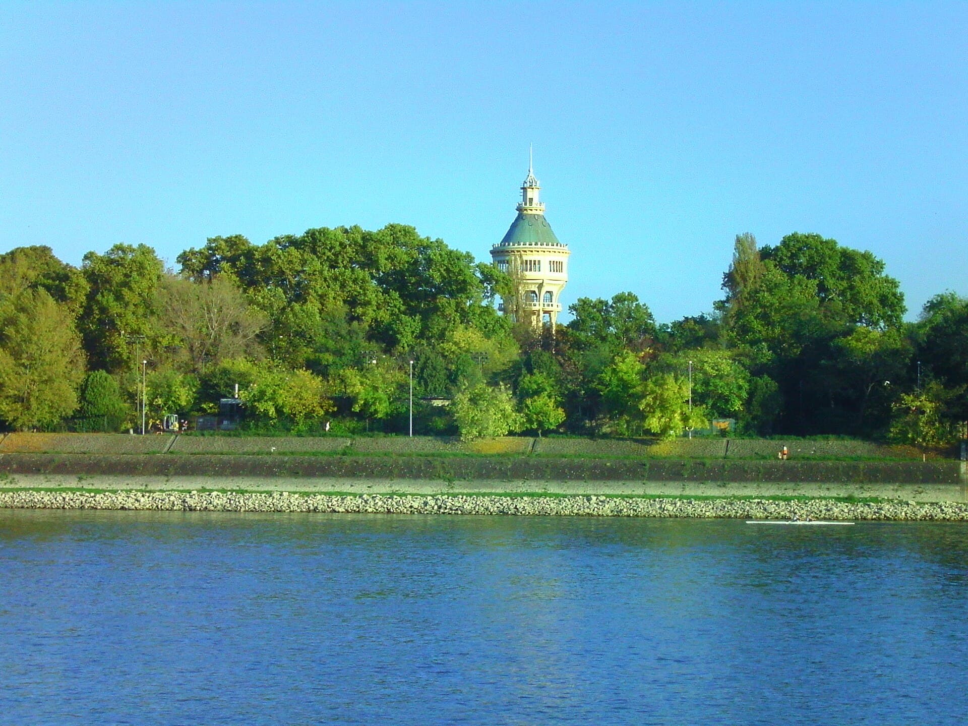 The Art Nouveau water tower of Margaret Island seen across the Danube, its octagonal lantern and green dome rising above a line of trees, with a single rowing boat on the water below