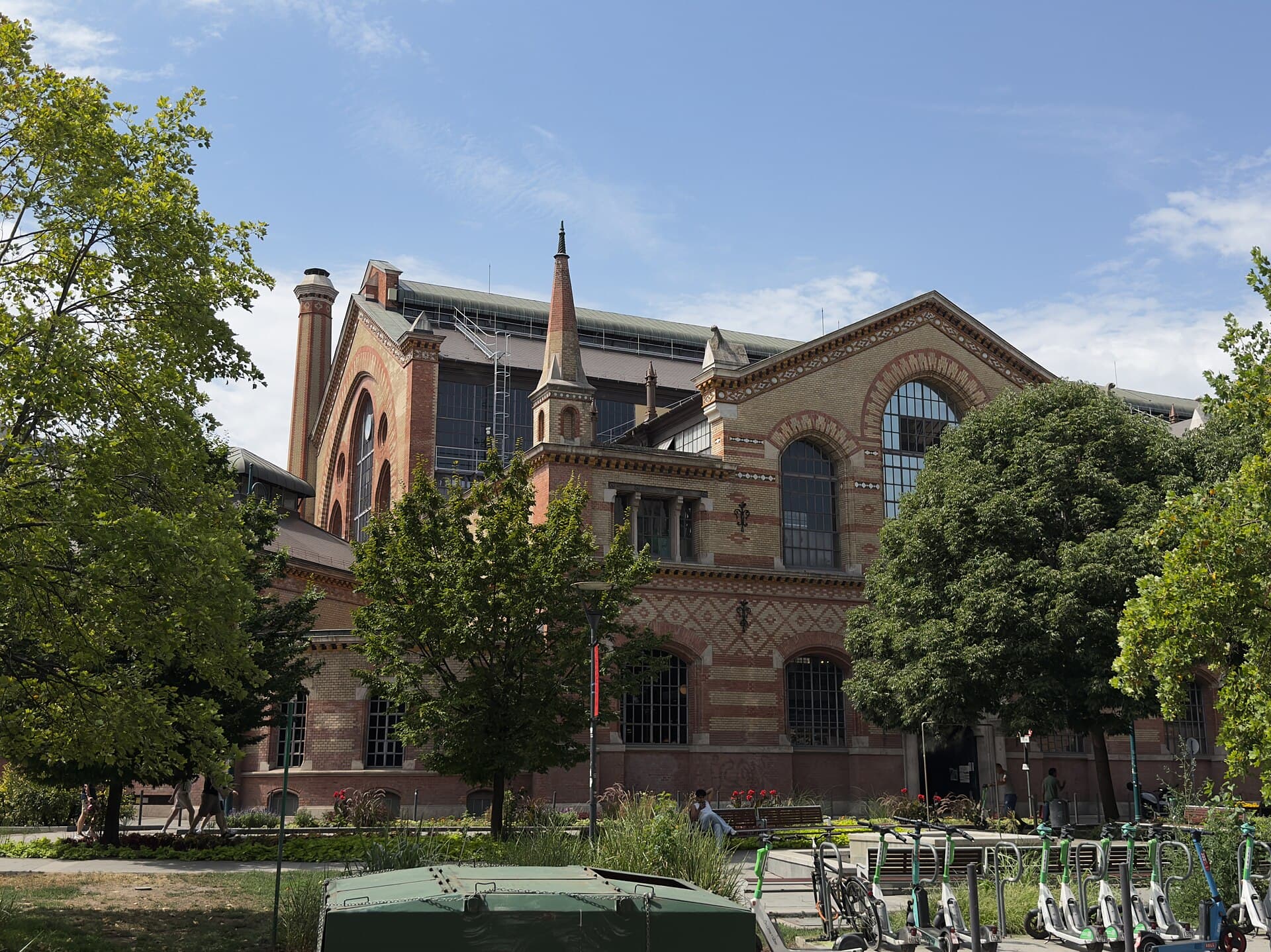 The brick and stone side elevation of the Great Market Hall in Budapest seen across a small park from Csarnok square, with tall arched windows, a slender spire, trees in front, and a rank of hire e-scooters parked along the pavement