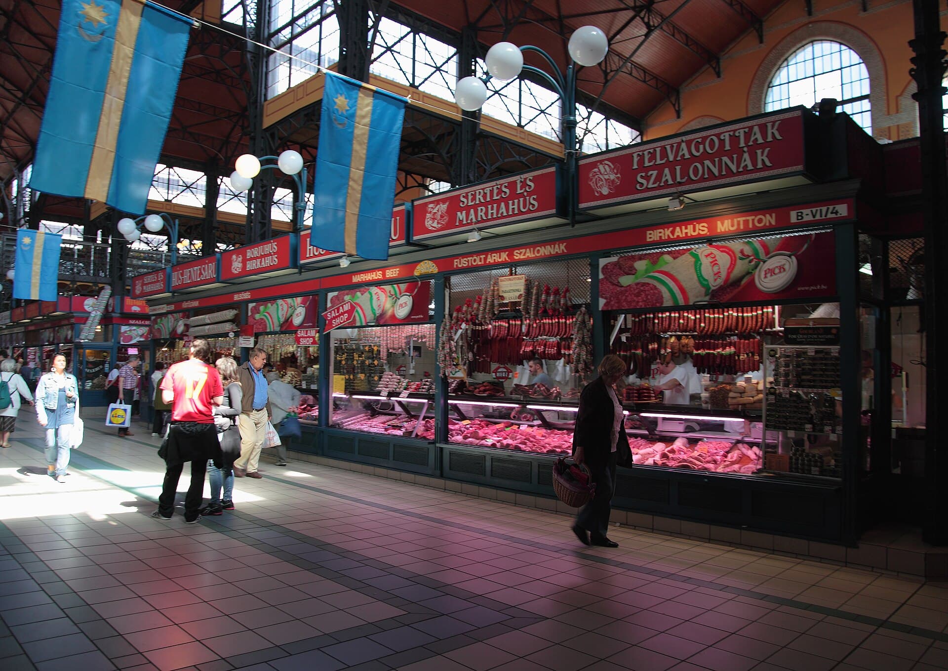 The ground floor of the Great Market Hall in Budapest: a long row of butcher counters under red signs reading SERTES ES MARHAHUS and FELVAGOTTAK SZALONNAK, strings of sausages hanging above chilled display cases, the steel roof structure overhead, and a woman walking past carrying a wicker shopping basket