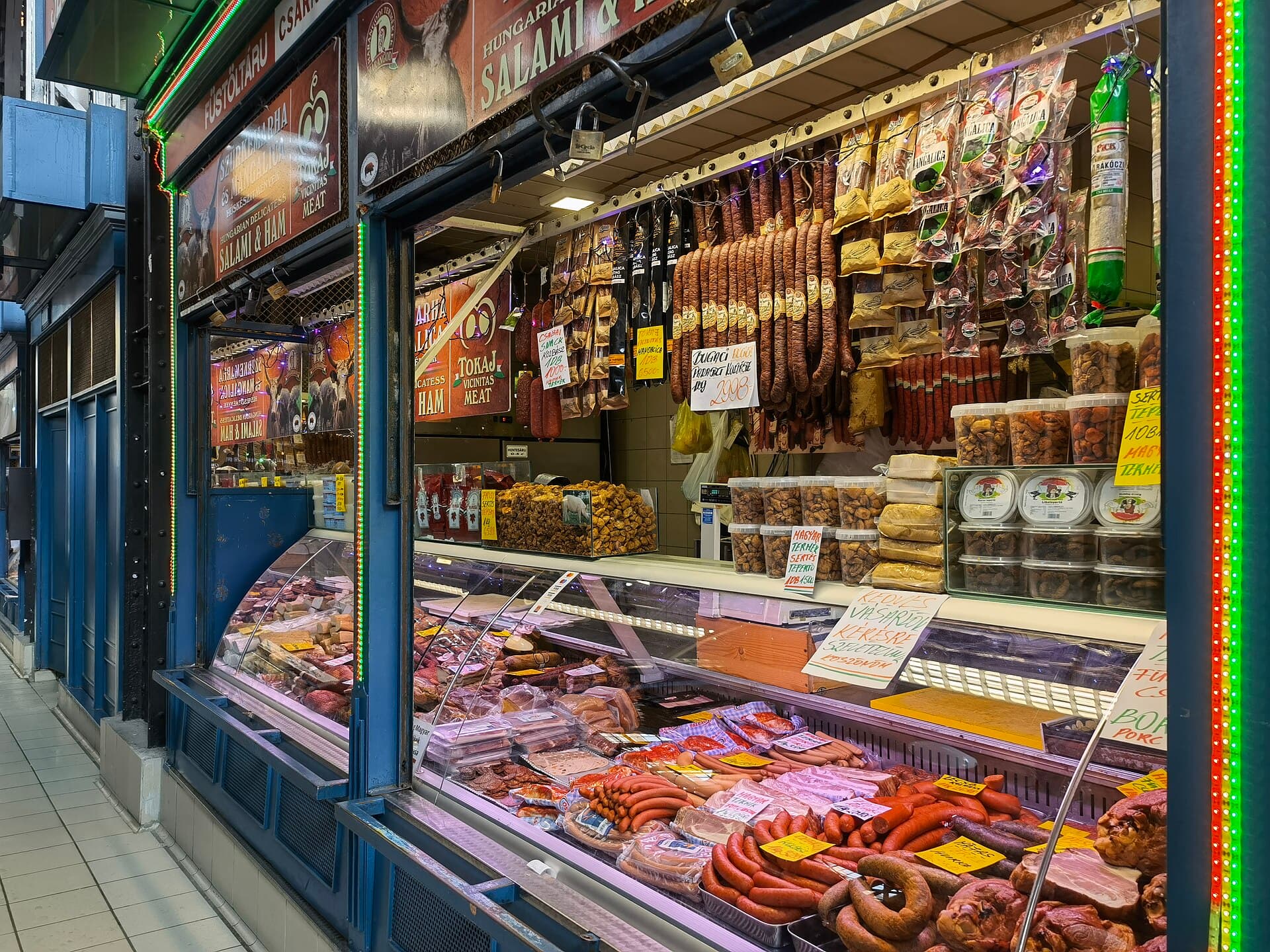 A salami and ham stall in the Great Market Hall with signs in English reading HUNGARIAN DELICATESS SALAMI AND HAM and TOKAJ VICINITAS MEAT, Mangalica-branded sausages hanging in packets above a chilled counter, tubs of pork scratchings, and handwritten price cards in forints