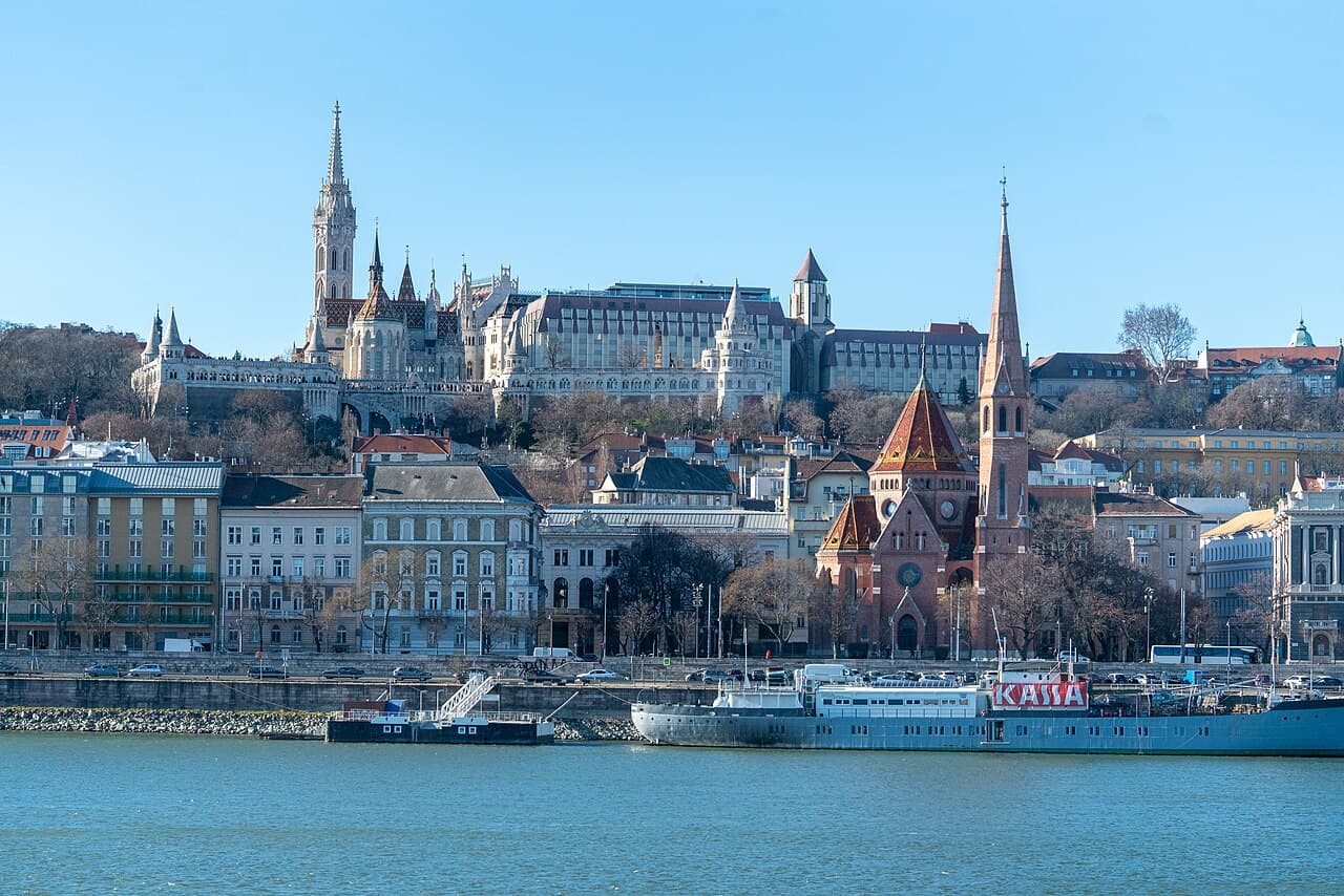 Castle Hill seen across the Danube from the Pest side on a clear winter day, with the white turrets and arcades of Fisherman's Bastion running along the hilltop and the spire and patterned roof of Matthias Church rising directly behind them