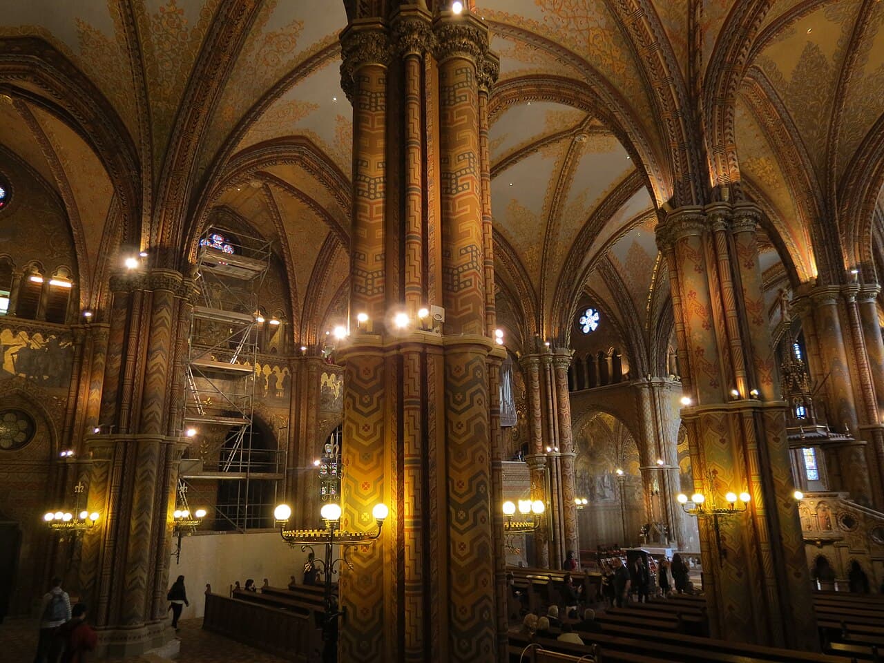 The interior of Matthias Church, with massive round columns and ribbed vaults covered from floor to ceiling in painted geometric patterns in ochre, gold and dark blue, wrought-iron candelabra holding white globe lamps, dark wooden pews, and restoration scaffolding standing against the left-hand wall