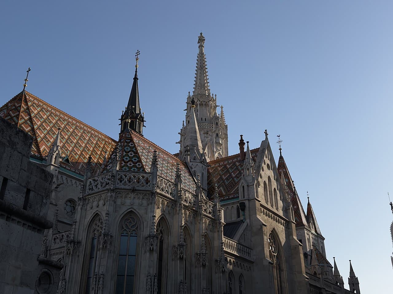 The diamond-patterned glazed tile roof of Matthias Church in orange, green and white, seen from below against a clear sky, with the pinnacled white stone spire rising behind it and a smaller dark spire topped by a gold cross on the left
