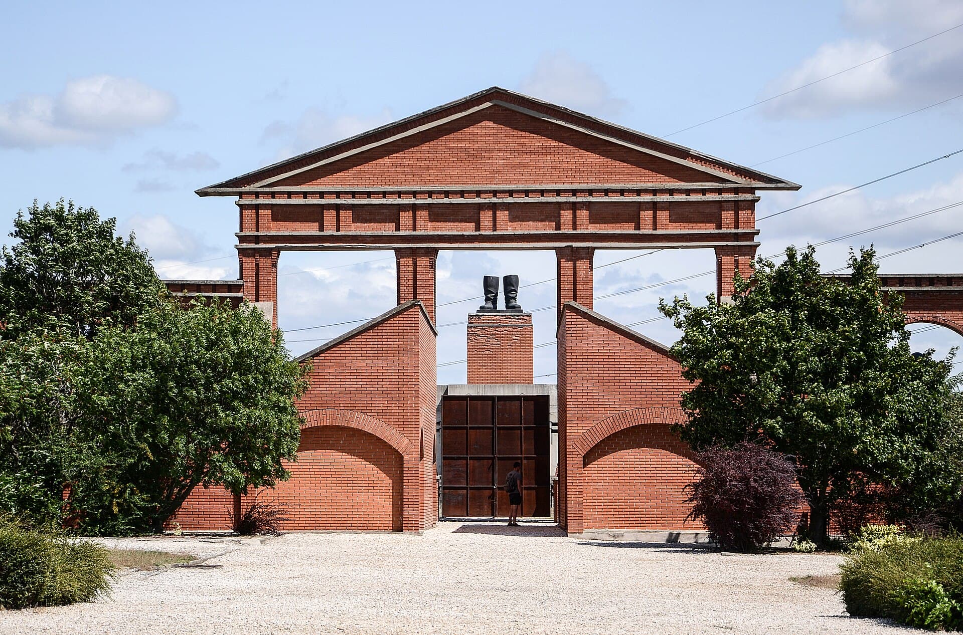 The red brick entrance facade of Memento Park, a classical pediment with no building behind it, with a pair of giant bronze boots standing on a plinth in the opening and a single visitor at the iron gate below