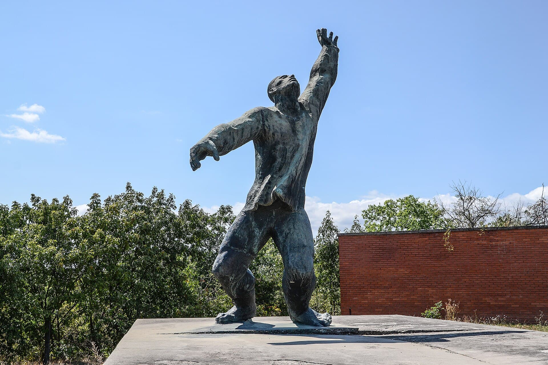 A large weathered bronze figure striding forward with one arm flung up towards the sky, standing on a bare concrete slab against a red brick wall and open countryside