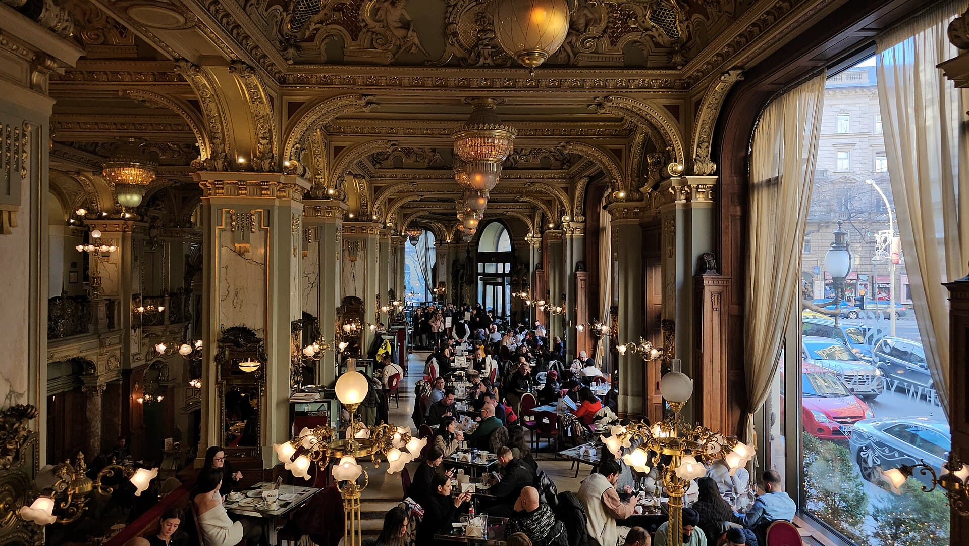 The same room at a busy hour: every table occupied, waiters moving between them, a line of people standing along the back wall waiting to be seated, and the traffic of the Grand Boulevard visible through a tall window on the right