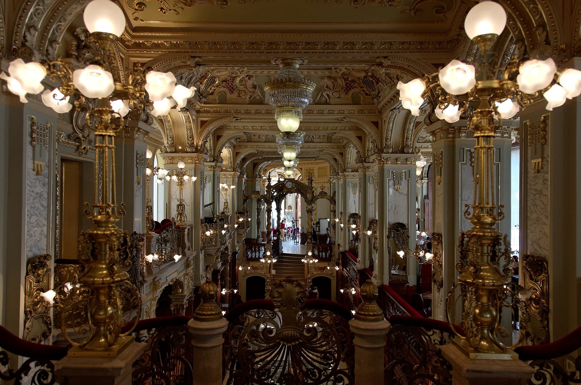 The interior of the New York Café in Budapest seen from the upper level: gilded candelabra with frosted glass globes in the foreground, spiralled marble columns, a heavily ornamented ceiling with frescoes and gold plasterwork, crystal chandeliers down the length of the room and a grand staircase at the far end