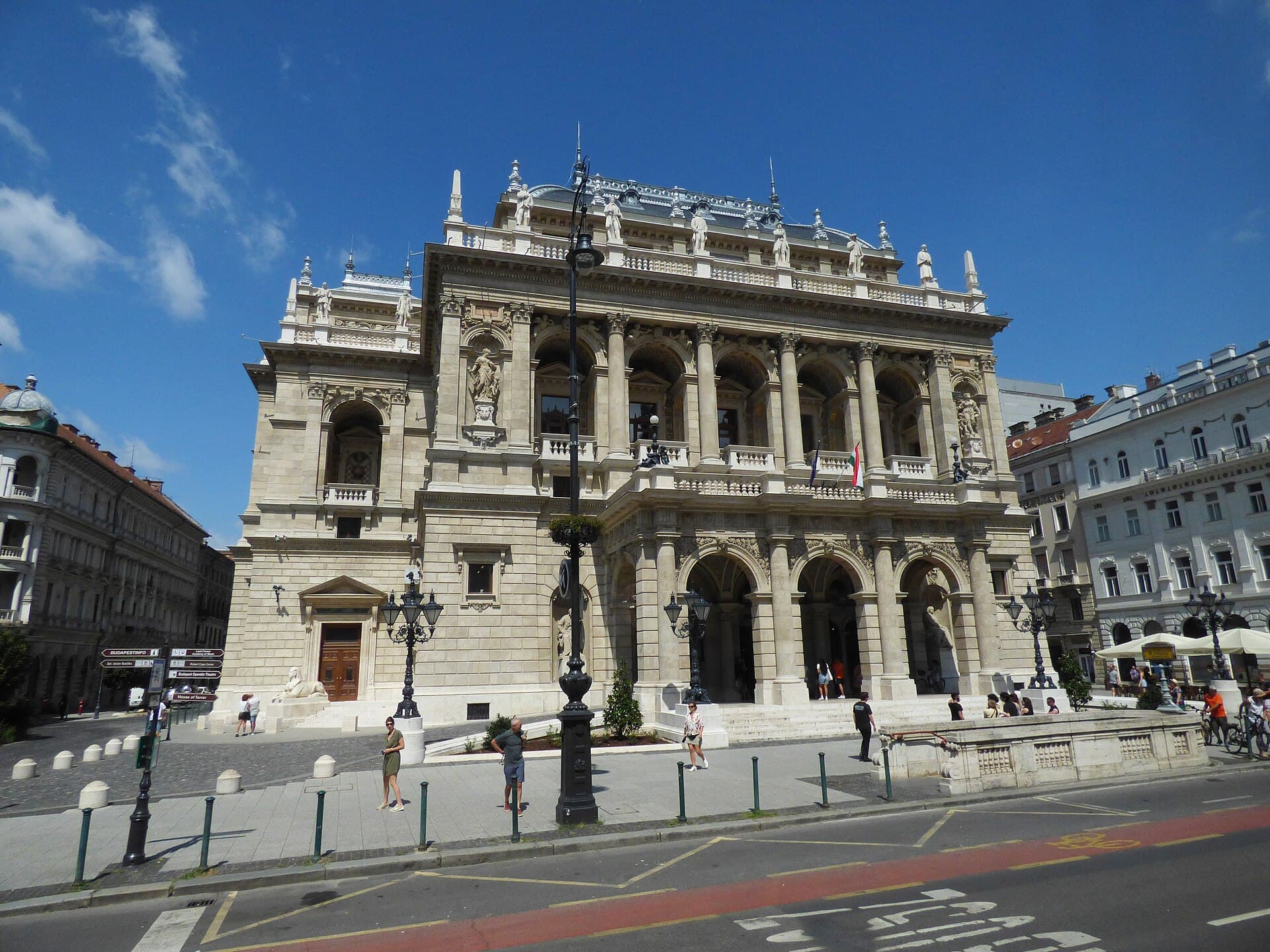 The Hungarian State Opera House on Andrassy Avenue in Budapest: a pale neo-Renaissance facade with an arcaded entrance at street level, a columned loggia above it, statues along the roofline and a sphinx beside the steps, with Hungarian and EU flags and people crossing the boulevard in front