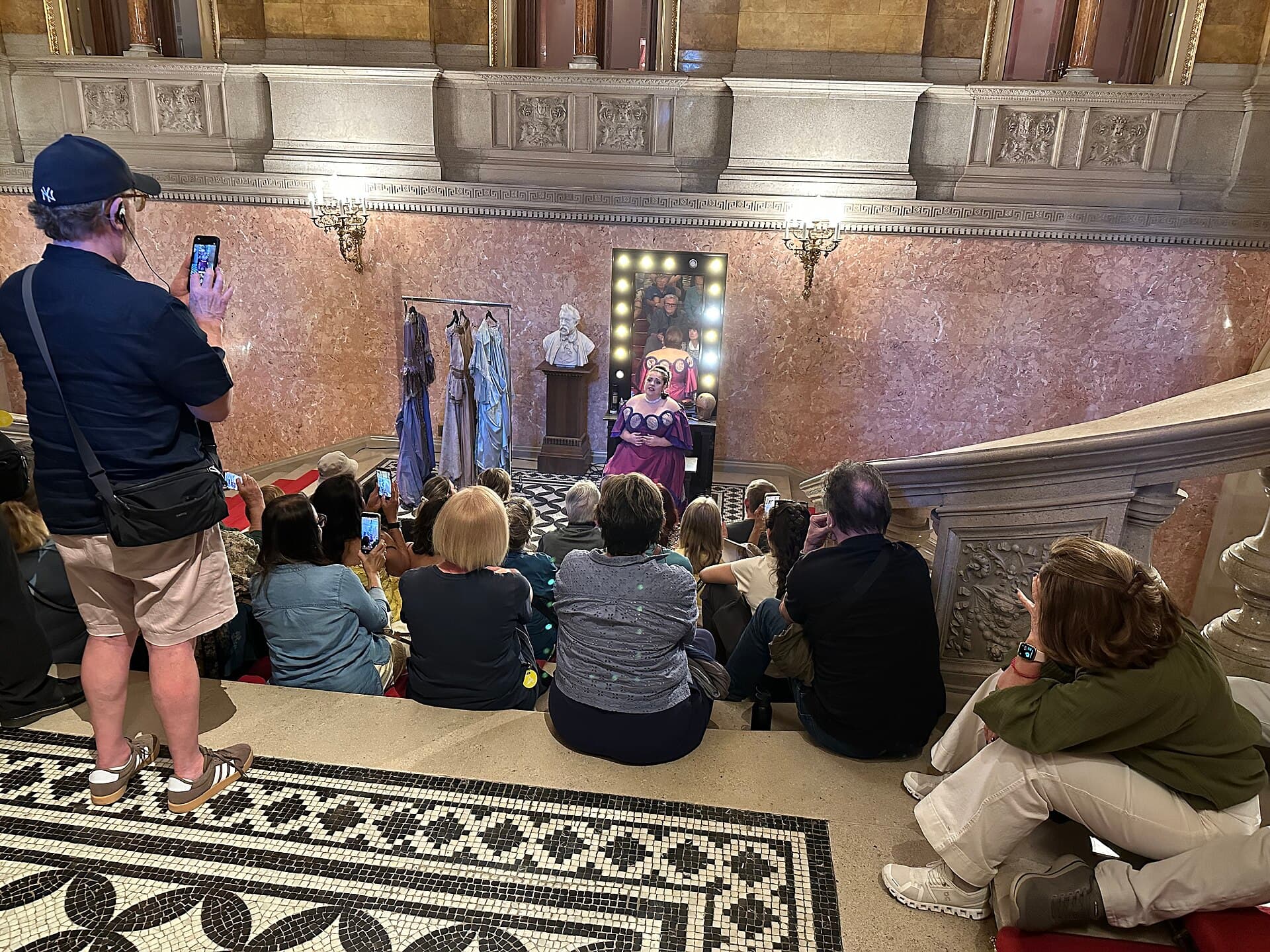 The short concert at the end of an Opera House tour: a soprano in a pink and purple costume singing on a landing beside a rail of costumes and a lit dressing-room mirror, watched by a tour group sitting on the marble steps, several of them filming on phones, with pink marble walls and a black and white mosaic floor
