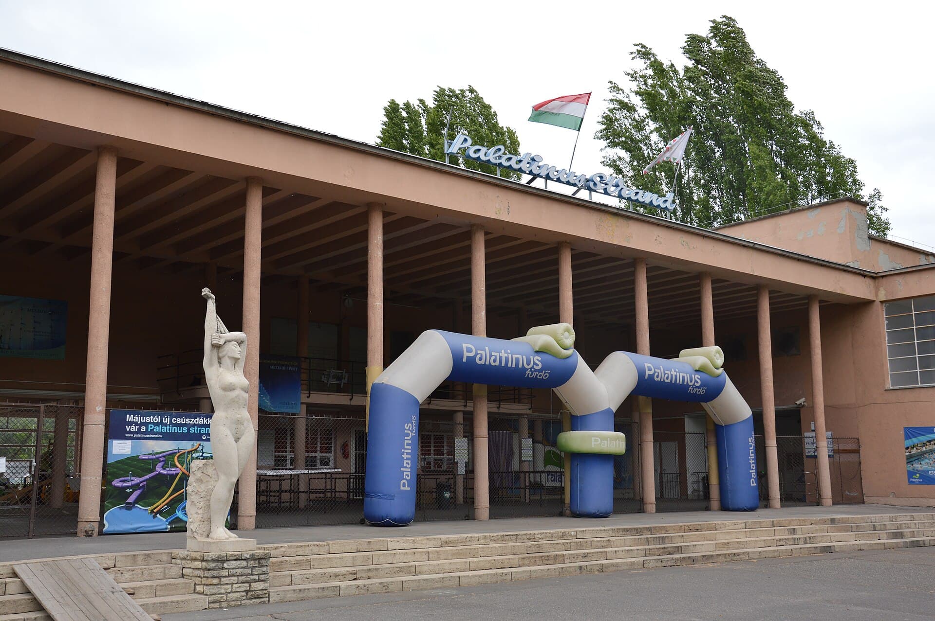 The long pink concrete colonnade of the Palatinus Strand entrance on Margaret Island, with the words Palatinus Strand in metal script on the roofline, a Hungarian flag flying above, two inflatable blue arches reading Palatinus furdo in front of the turnstiles and a white stone statue of a woman raising a towel beside the steps