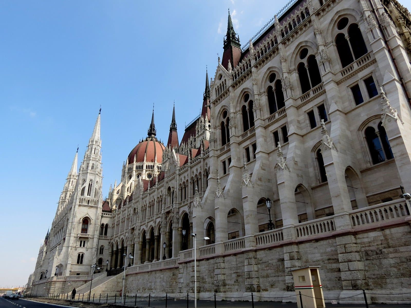 The Danube facade of the Hungarian Parliament seen from street level at a steep angle, showing the length of the pale stone frontage with its arcades, buttresses and spires receding into the distance, the red dome behind, and a small sentry box at the corner