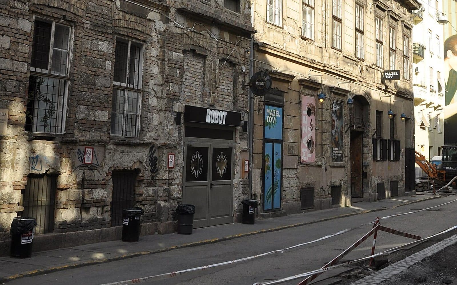 The shuttered grey doors of the ROBOT venue under its black sign on Akacfa utca in Budapest, in a building whose render has fallen away to bare brick, with the painted Mazel Tov panels on the next building along