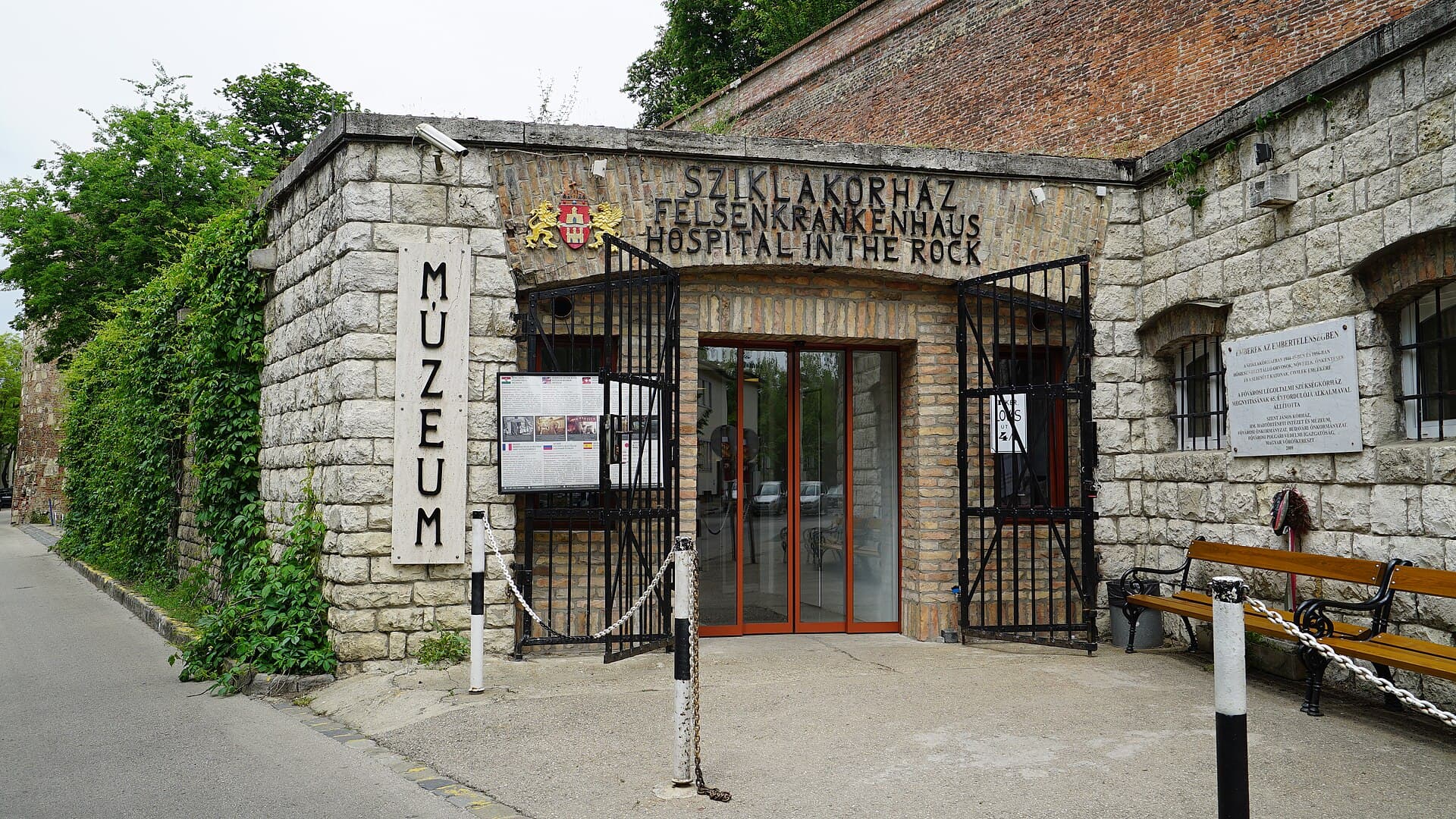 The entrance to the Hospital in the Rock museum in Budapest: a small stone doorway set into the castle wall with SZIKLAKORHAZ, FELSENKRANKENHAUS and HOSPITAL IN THE ROCK carved above it in three languages, a vertical MUZEUM sign to the left, open iron gates and red-framed glass doors