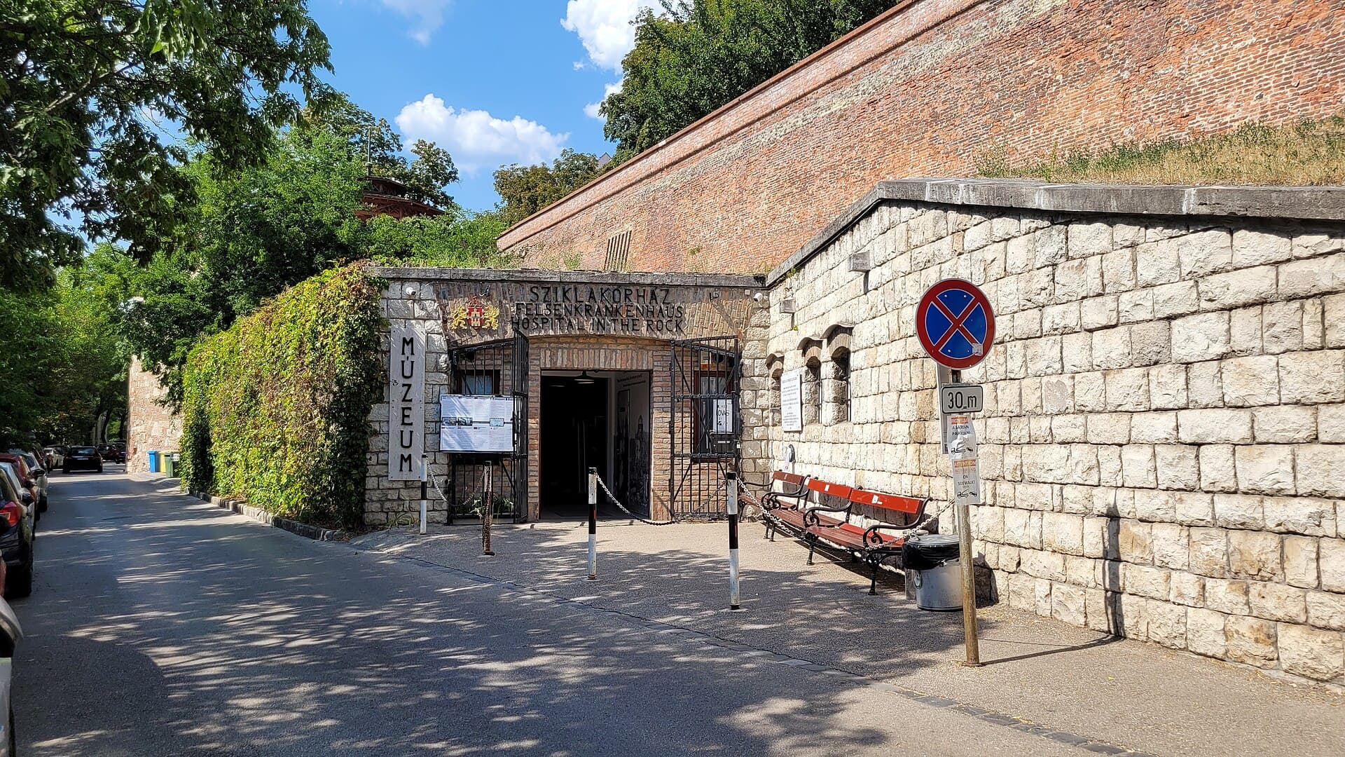Lovas Street running along the foot of the Buda Castle wall in summer, with the small stone entrance to the Hospital in the Rock set into the wall on the right, the brick fortification rising above it, a bench outside and cars parked along the kerb