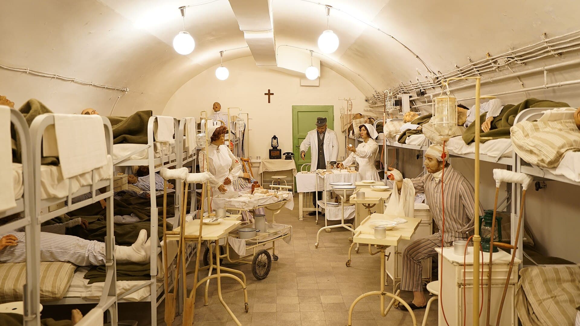 A reconstructed hospital ward inside the Hospital in the Rock: a long white vaulted tunnel lined with metal bunk beds holding wax figures of wounded patients under green blankets, nurses in white uniforms and a doctor in a white coat and cap tending to them, drip stands, crutches and enamel bowls between the beds, and a crucifix on the far wall