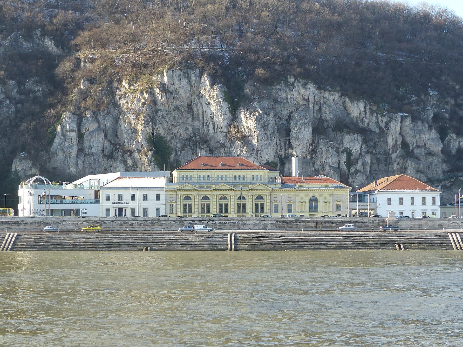 The Rudas bath complex seen from across the Danube in winter, with the yellow neoclassical block in the middle, the Rudas Restaurant signage on the left and the ribbed white dome of the rooftop pool above it, all under the cliffs of Gellert Hill
