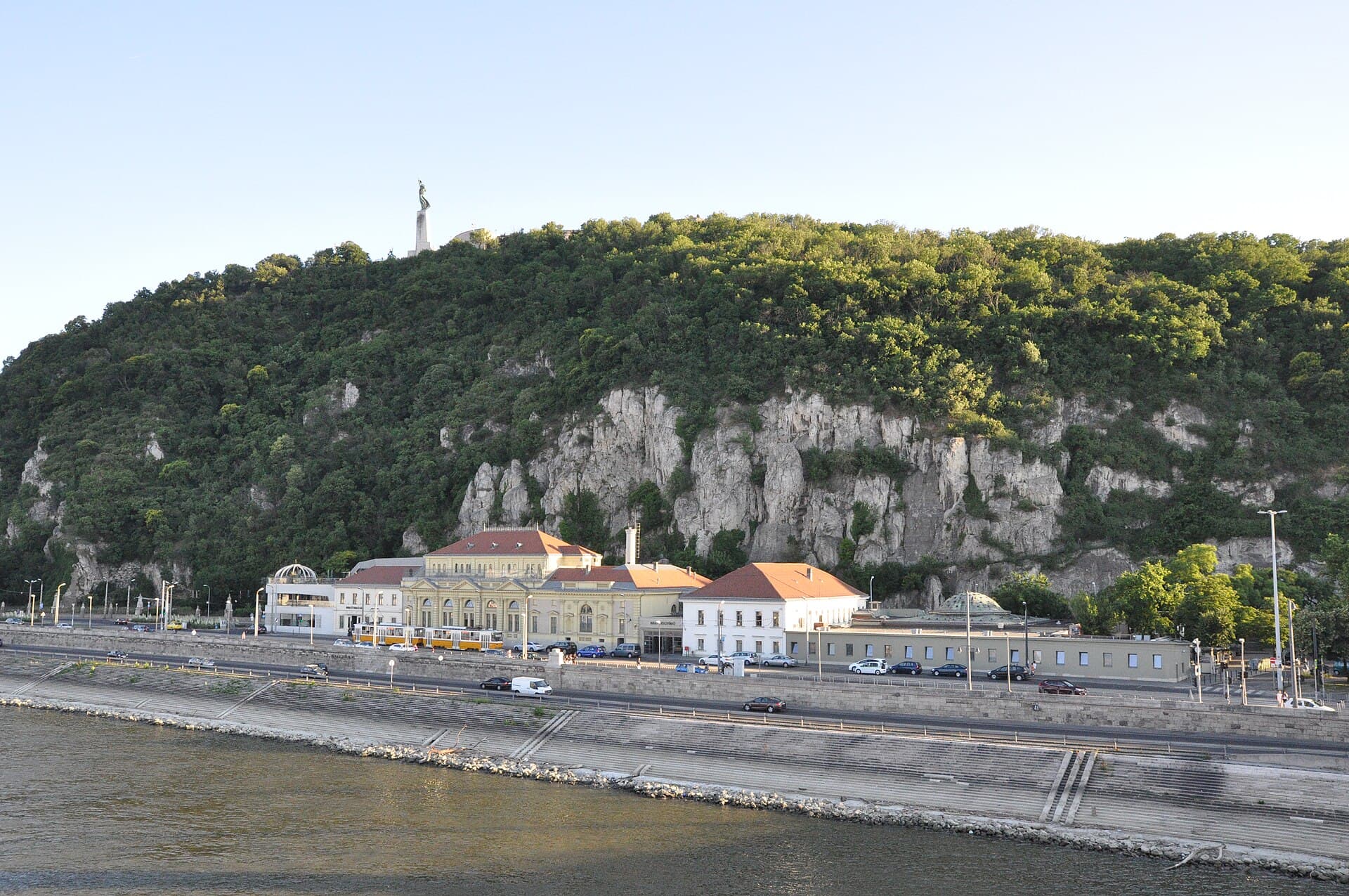 Rudas seen from across the Danube in summer, a low cluster of buildings pressed against the wooded cliffs of Gellert Hill with the Liberty Statue on the summit above and a yellow tram passing on the embankment