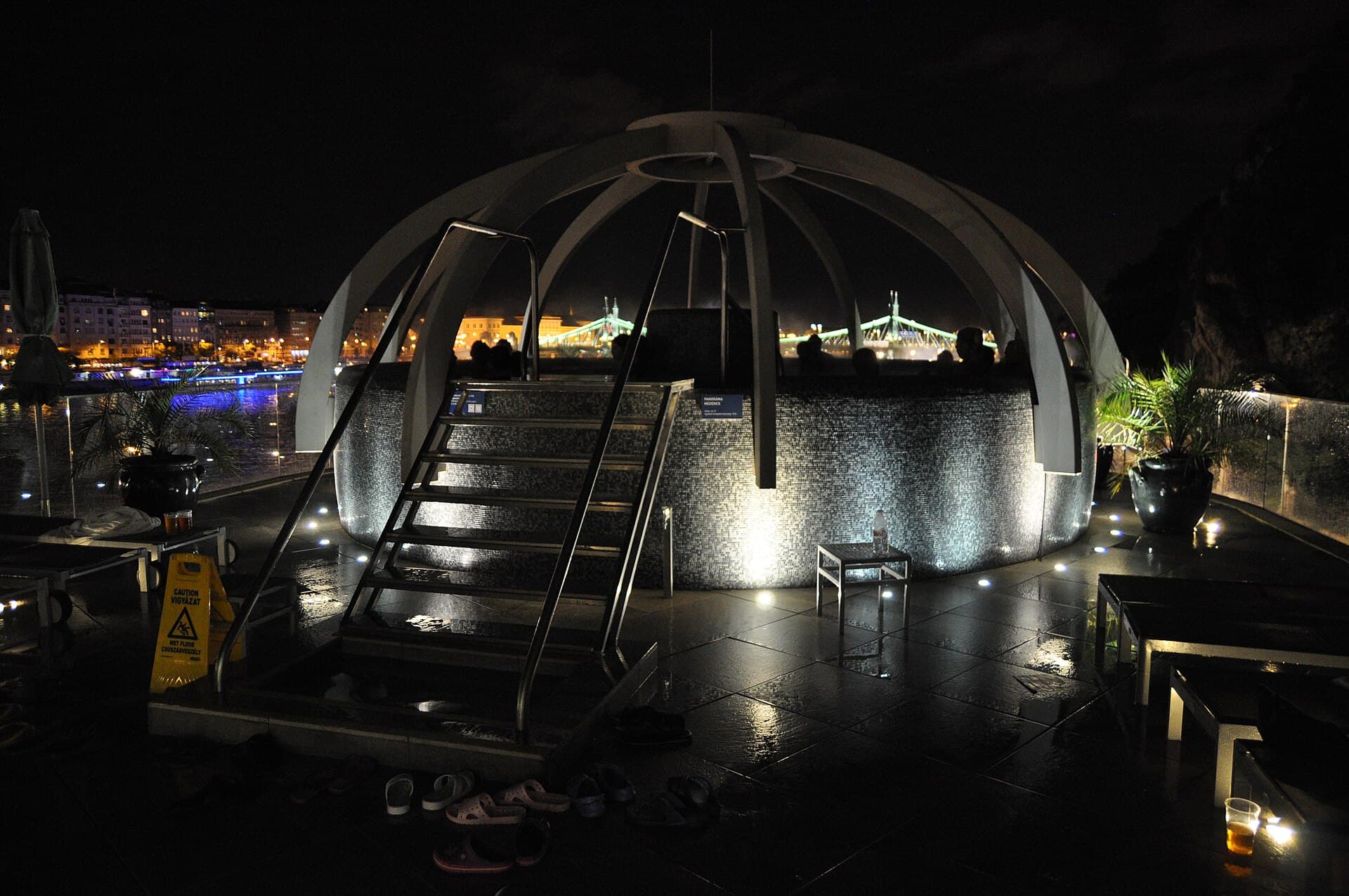 The rooftop panorama pool at Rudas at night, a circular pool under a ribbed open dome with bathers leaning on the far rail, the lit Liberty Bridge across the Danube behind, and sandals left on the wet tiles in the foreground