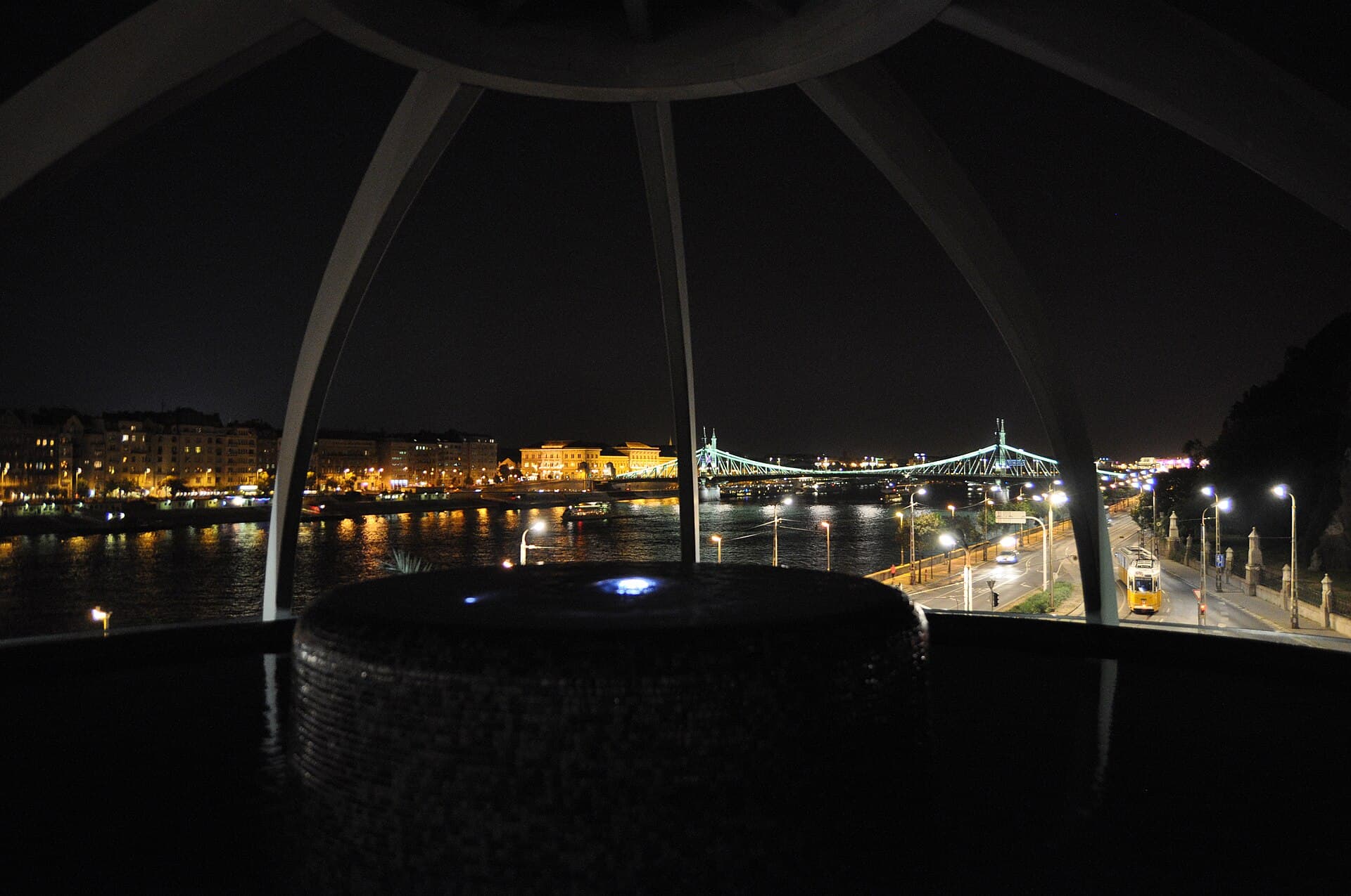 The view out from inside the rooftop pool at Rudas after dark, framed by the ribs of the dome, showing the Danube, the lit Liberty Bridge and a yellow tram running along the embankment below