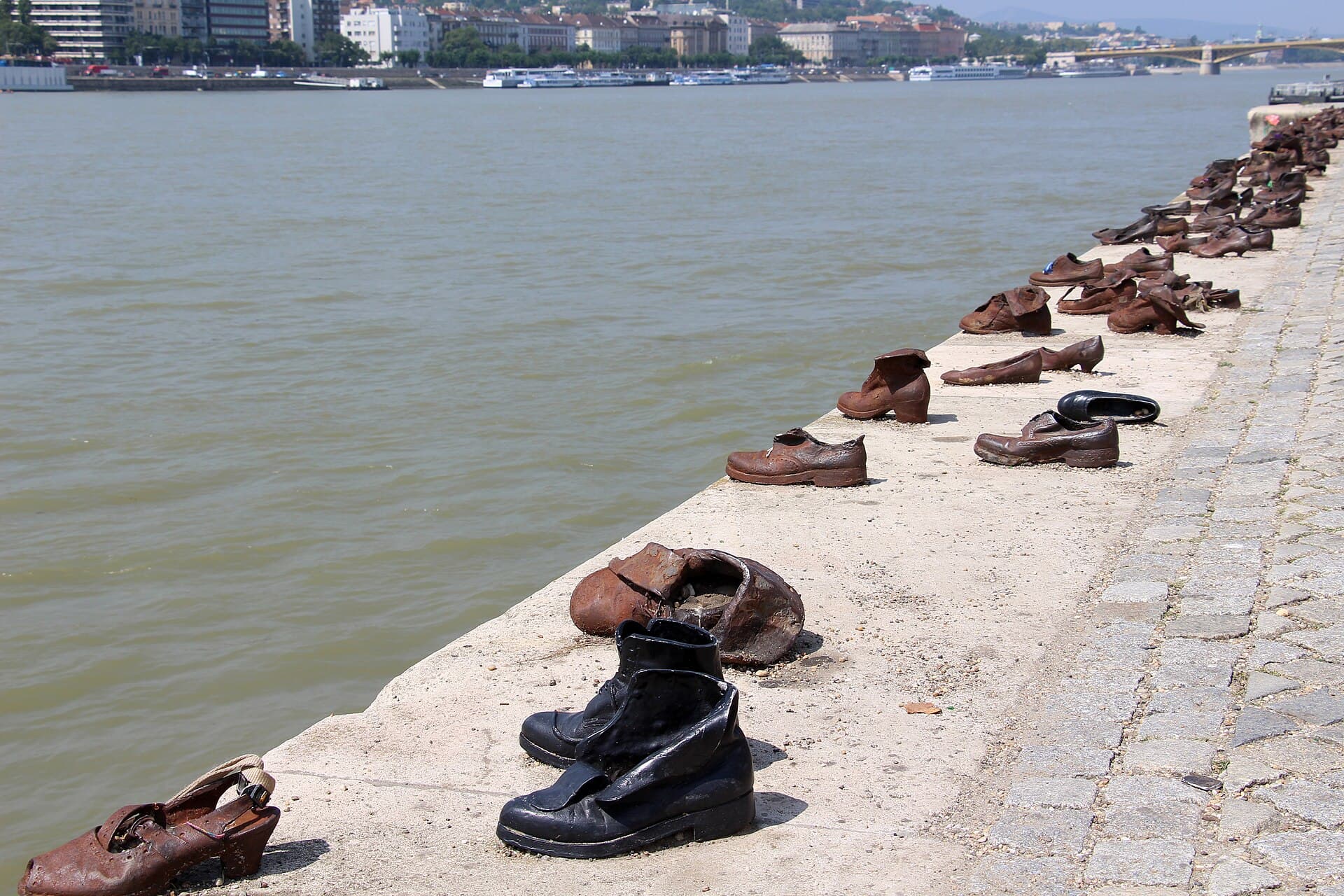 A closer view along the memorial showing individual rusted iron shoes on the concrete edge of the embankment: a child's small buckled sandal in the foreground, a heavy black work boot, a woman's heeled shoe, and small stones left inside some of them, with the Danube alongside