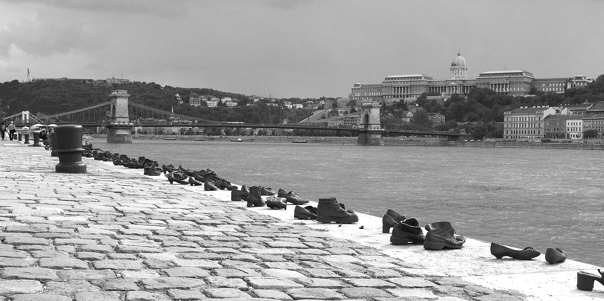 A black and white photograph of the Shoes on the Danube memorial: a long line of iron shoes set into the edge of the stone embankment, running away into the distance beside the water, with the Chain Bridge and Buda Castle on the far bank under an overcast sky