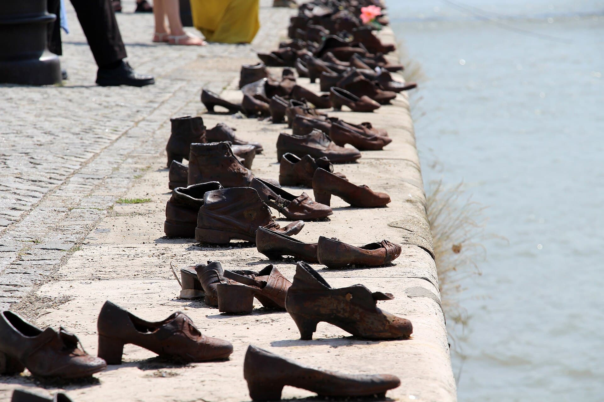 Visitors' feet in sandals and dress shoes at the top of the frame as they walk along the promenade past the line of rusted iron shoes set into the embankment edge, with a single pink flower left in one of the iron shoes further along
