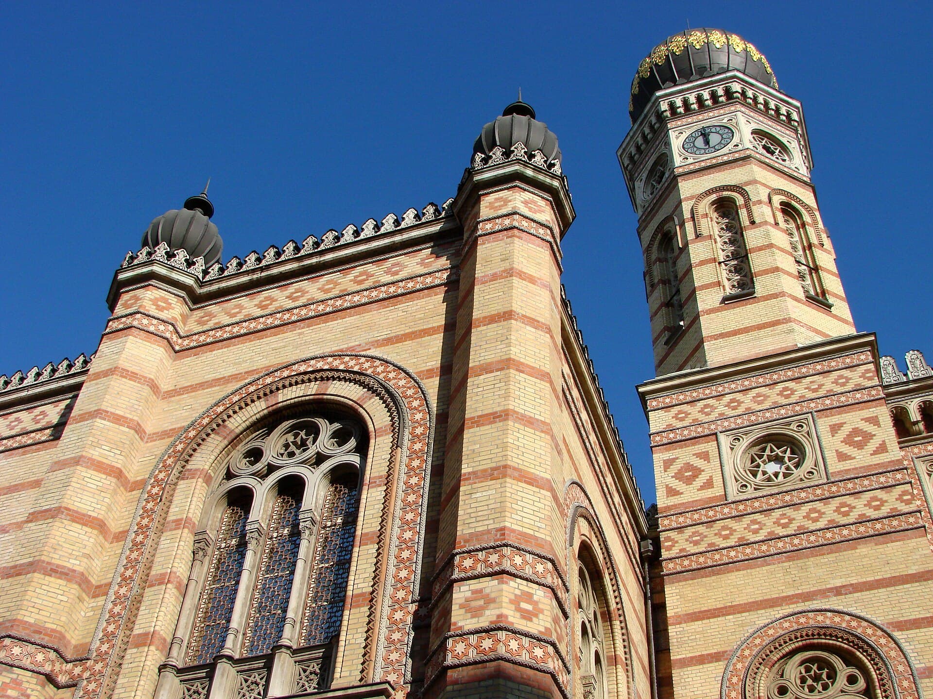 The Moorish Revival facade of the Dohány Street Synagogue against a deep blue sky, showing the striped yellow and red brickwork, the star-patterned rose window and one of the onion-domed clock towers