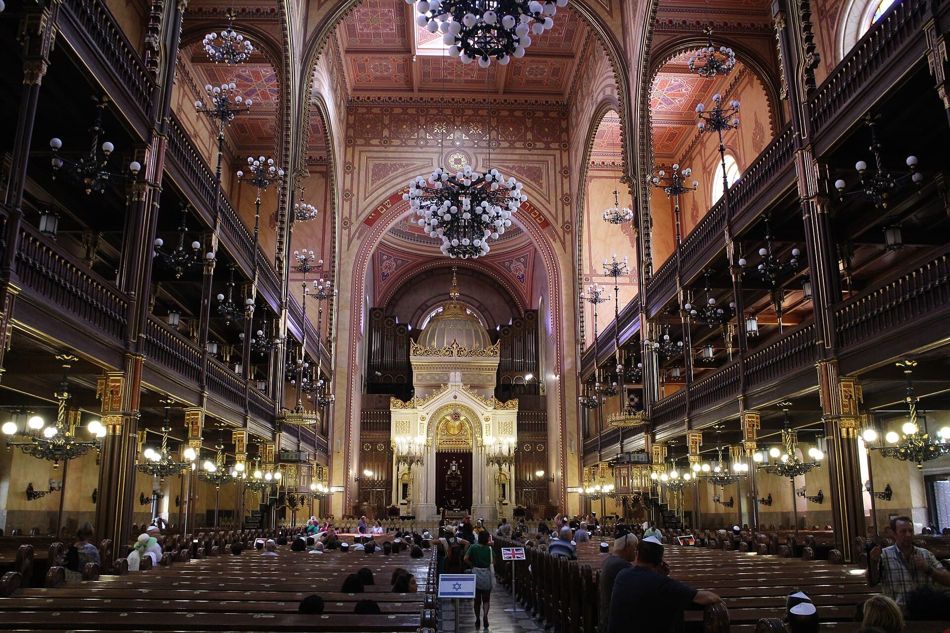 The interior of the Dohany Street Synagogue looking down the aisle towards the ark and organ, with two tiers of galleries, rows of chandeliers, and visitors seated in the pews beside small national flags on stands