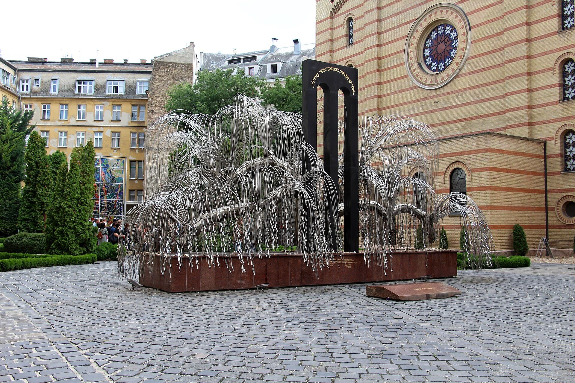 The Emanuel Tree of Life memorial in the Raoul Wallenberg Memorial Park: a metal weeping willow whose leaves are inscribed, standing on a red granite base in a cobbled courtyard against the striped brick wall of the synagogue