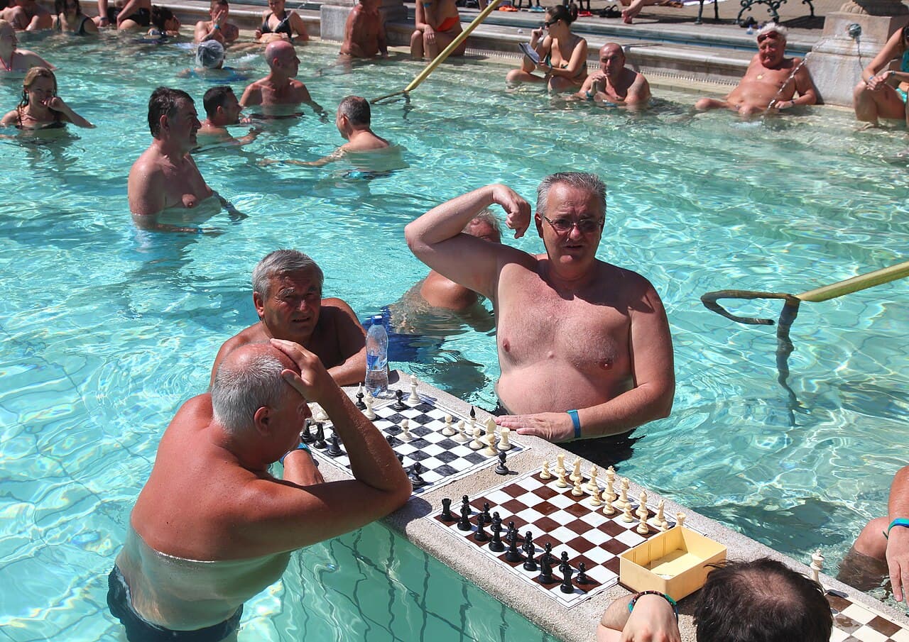 Varios hombres mayores metidos en el agua hasta el pecho en la piscina exterior del Széchenyi, jugando al ajedrez sobre dos tableros apoyados en una repisa de piedra que cruza la piscina, con más bañistas al fondo