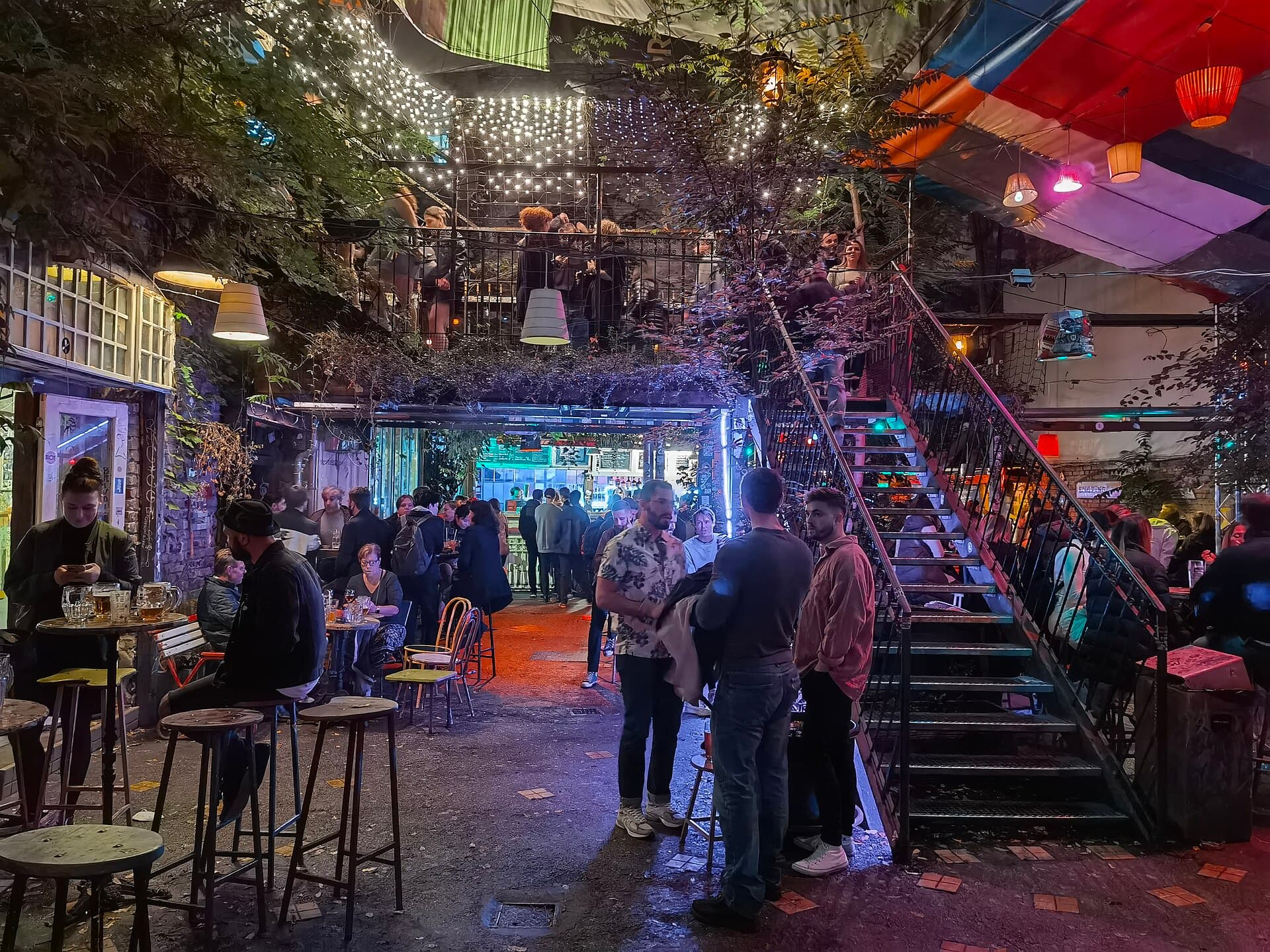 El patio principal del Szimpla Kert de noche, lleno de gente bebiendo bajo guirnaldas de bombillas, con una escalera metálica que sube a una galería abarrotada en el primer piso